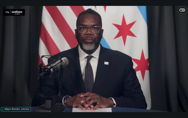 Chicago Mayor Brandon Johnson stands at a podium behind a microphone, and in front of the Chicago flag.