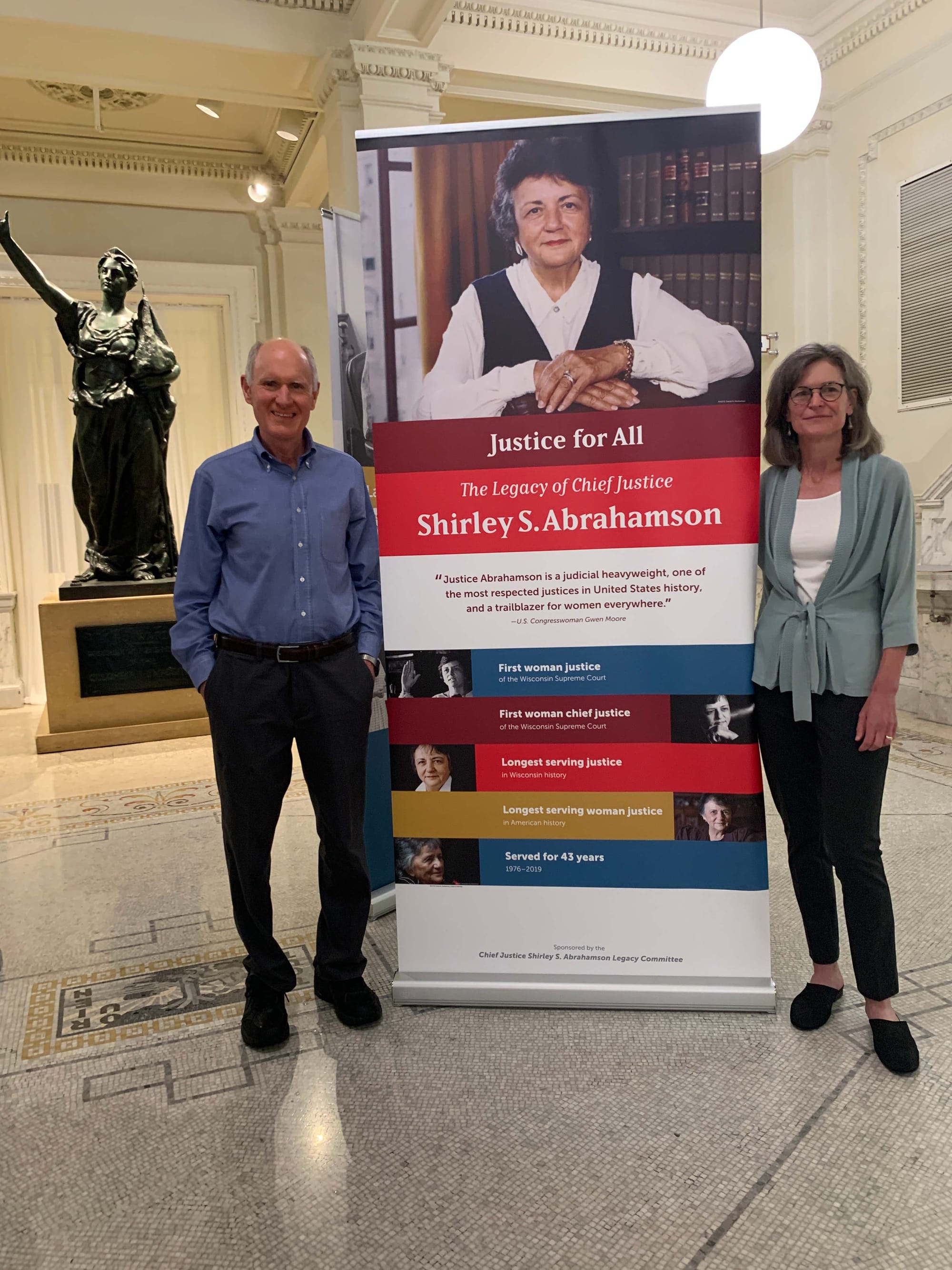 Colleen Ball and her husband stand on either side of a tall banner that features a portrait of Shirley Abrahamson above the title Justice for All: The Legacy of Chief Justice Shirley S. Abrahamson