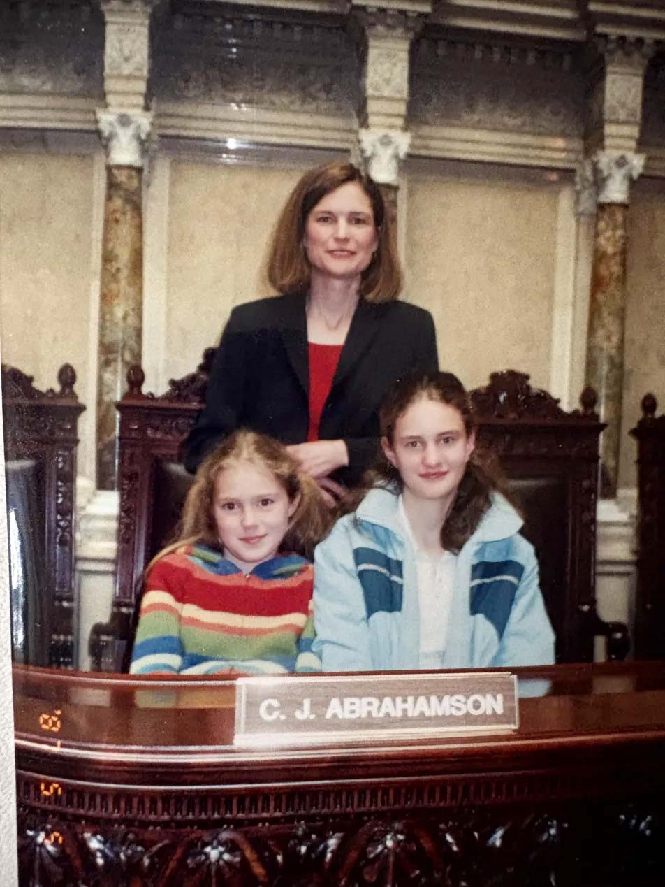 Colleen Ball in a red top and black blazer stands behind two young girls who are sitting behind a nameplate that reads "C. J. Abrahamson"