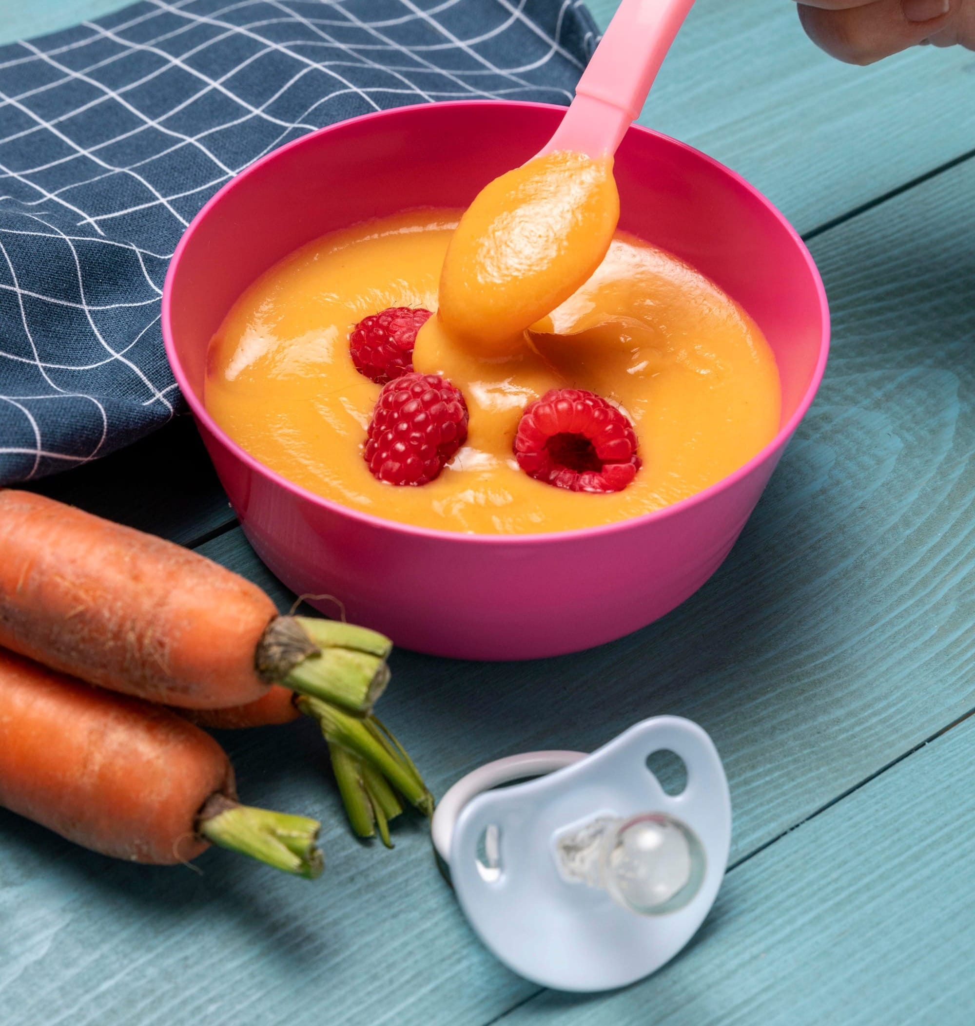 Bol de purée de carotte avec framboises fraîches, tenu par une main, posé sur une table en bois avec carottes et tétine à côté