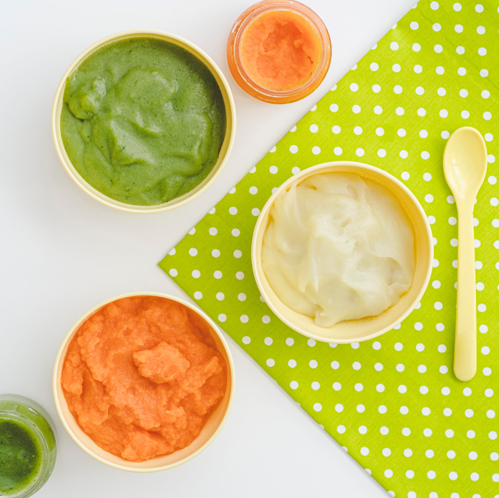Assortiment de purées pour bébé maison dans des bols jaunes : purée de carotte, courgette et pomme de terre, avec cuillère et serviette verte à pois