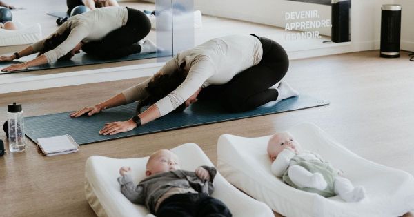 Une maman en posture de yoga postnatal pendant qu’un groupe de bébés repose sur des matelas ergonomiques dans une salle dédiée au bien-être chez Yada.