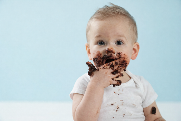 Bébé d’environ un an avec le visage et les mains couverts de mousse au chocolat, dégustant un dessert fait maison, en format paysage sur fond bleu clair.