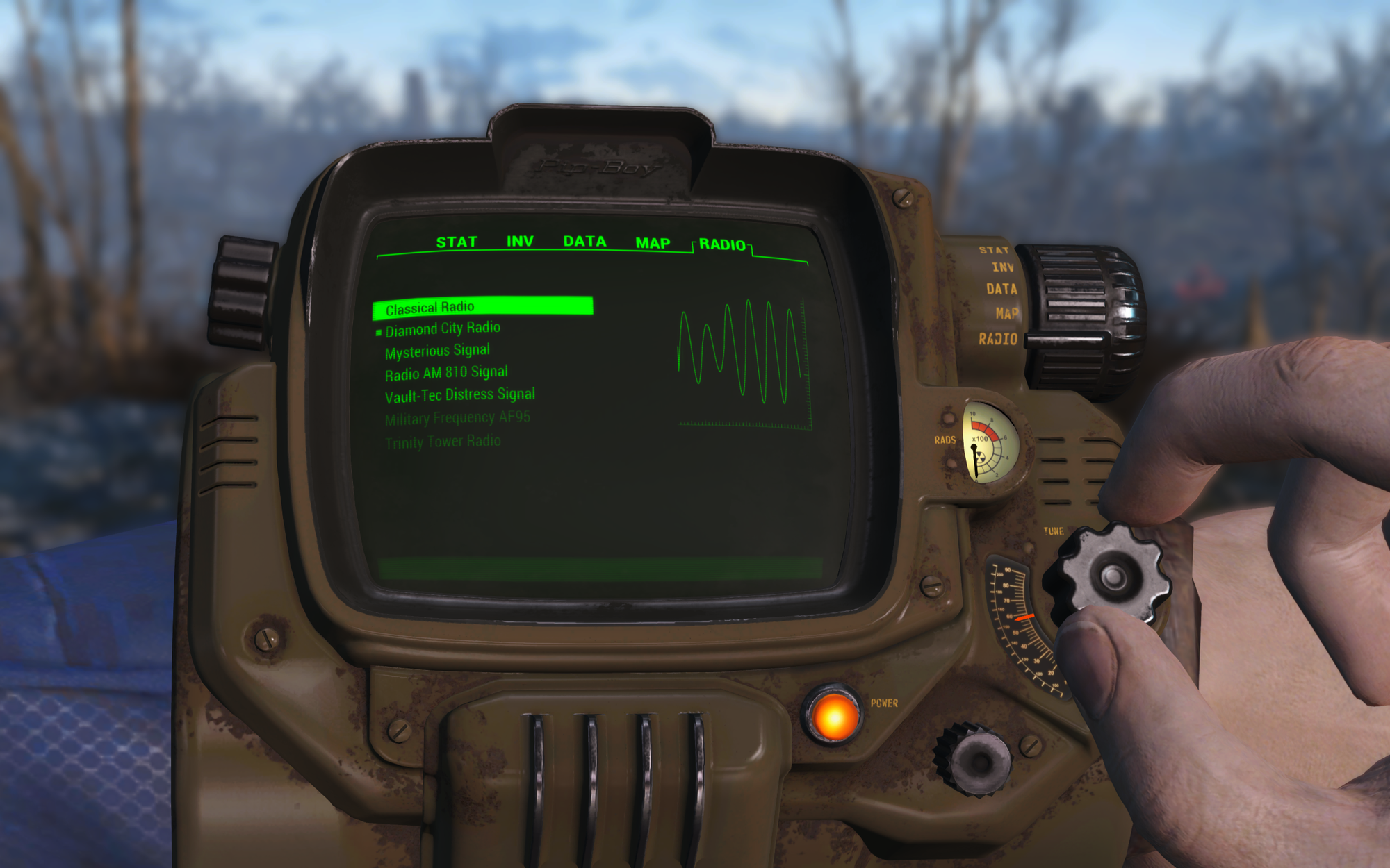 A screenshot from Fallout 4, the player's arm has a Pip-Boy screen showing radio stations. It is tuned into Diamond City Radio.