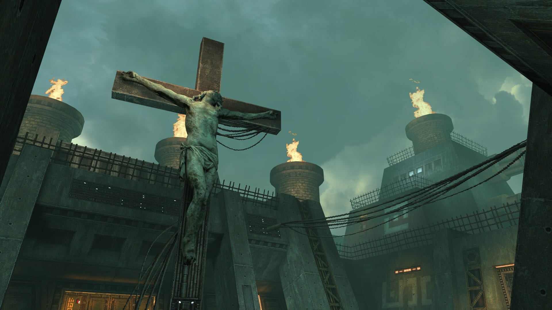 A crucifix in the centre of a concrete courtyard, with flaming towers in the background.