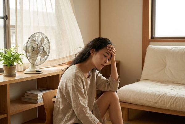 一名女子在日式木質房間裡，坐在電風扇旁扶著額頭，神情疲憊