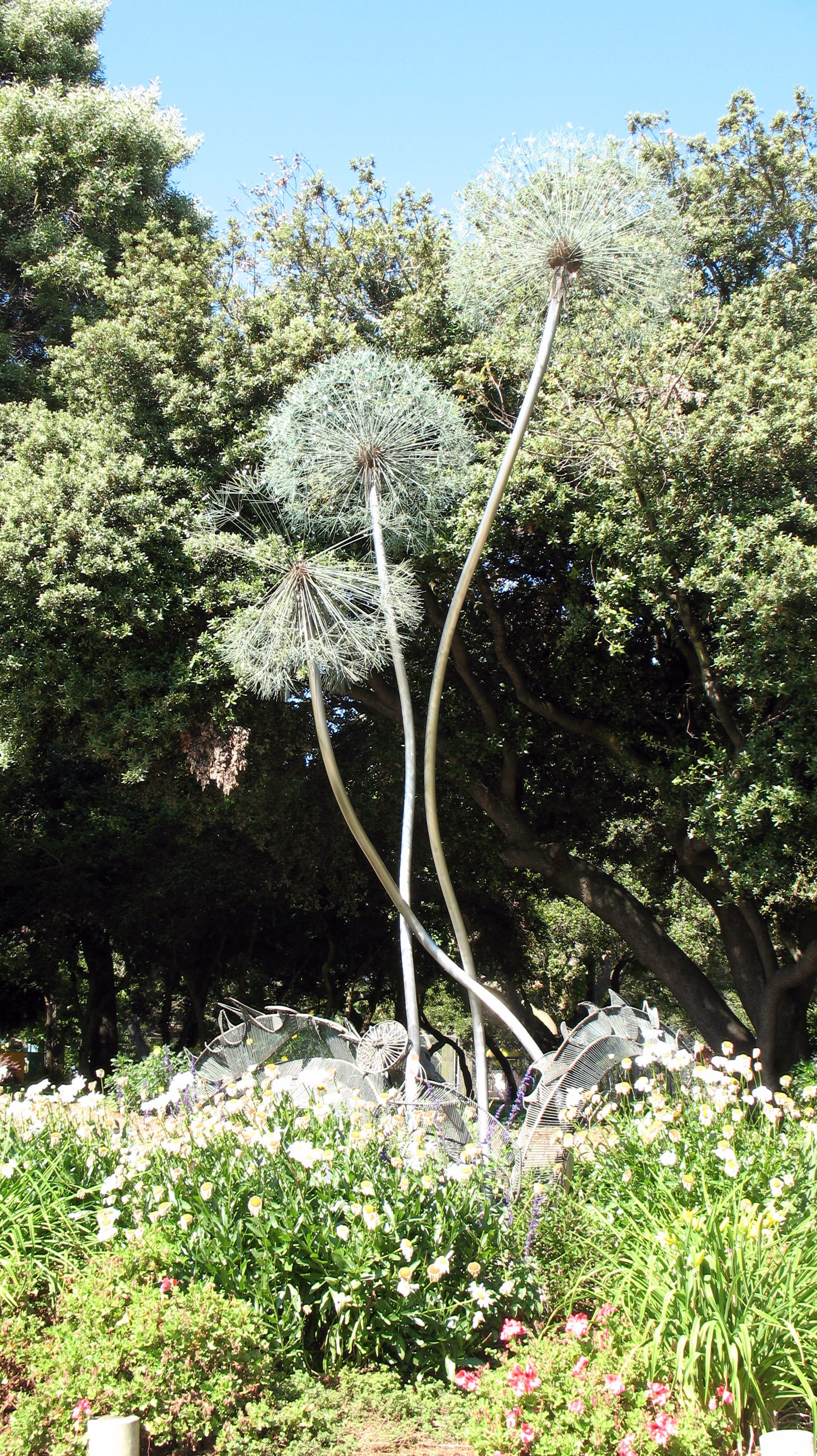 Three dandelion puffs made of metal, reaching into the sky. In the background are tall oaks, and in the foreground are flowers. One of the dandelion puffs looks a little worse for wear.
