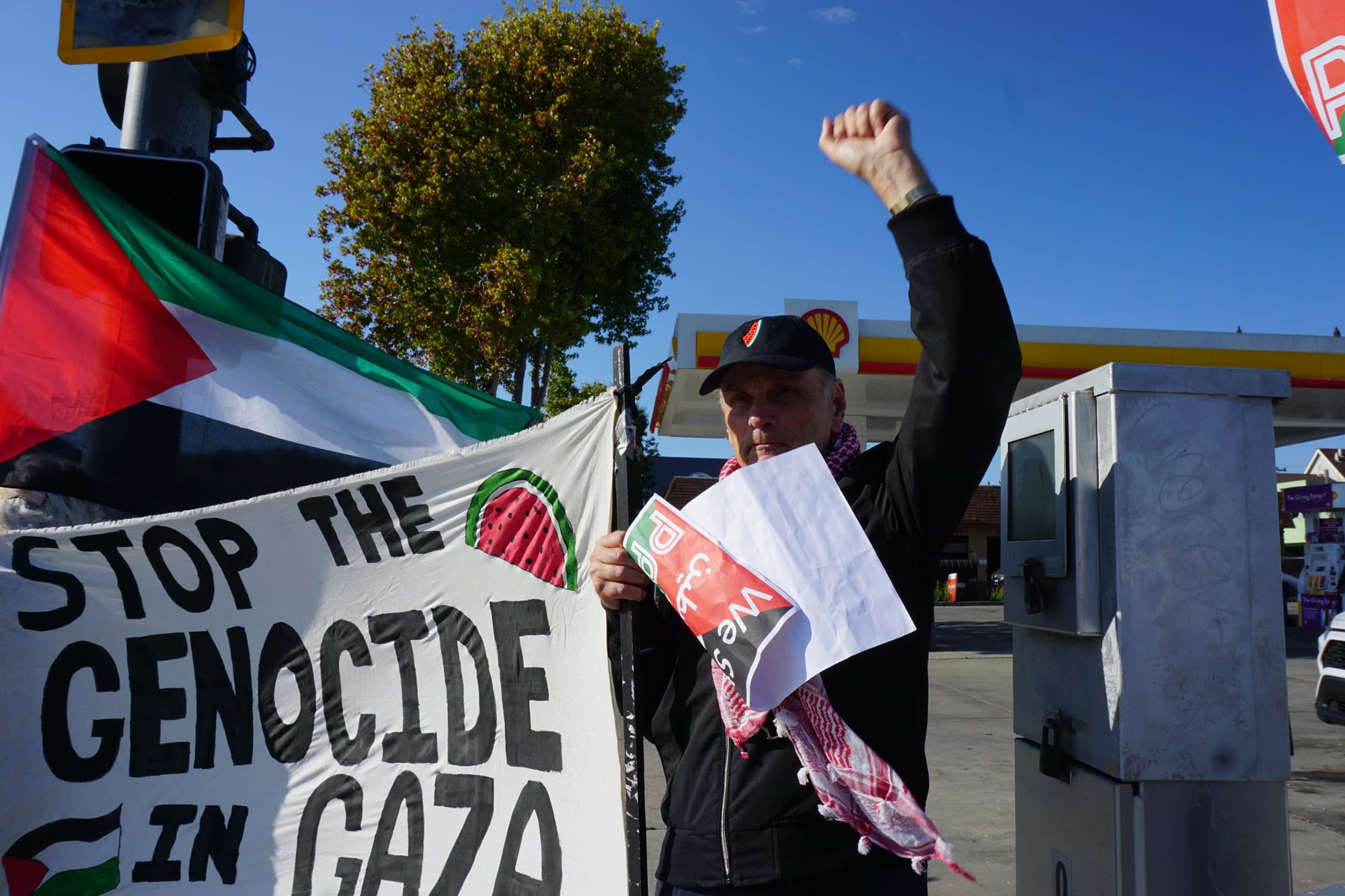 a man in his 50s holds a fist in the air while standing in front of a gas station, holding a sign that reads 'stop the genocide in gaza'