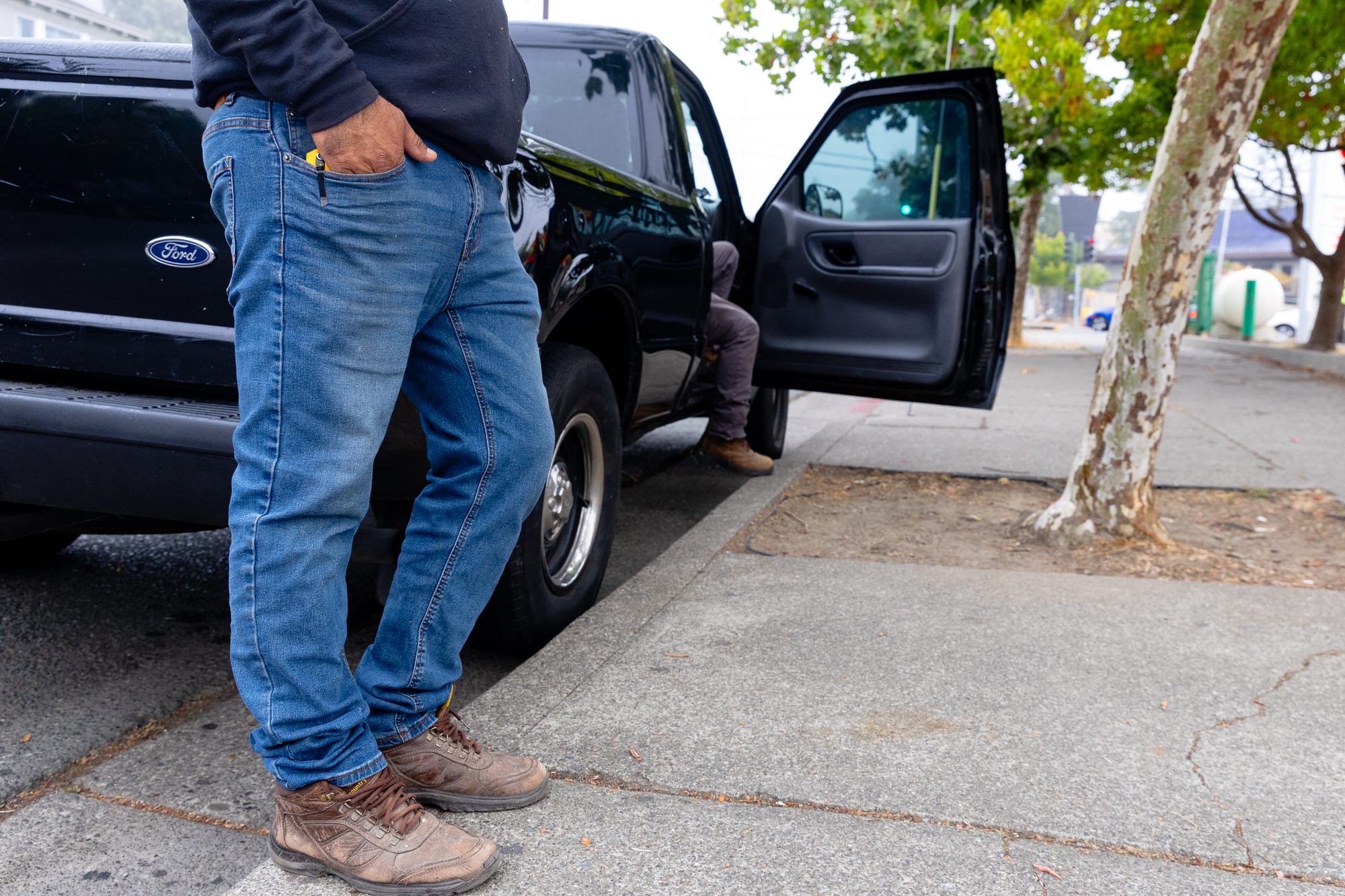 a man is shot from the waist down standing on the street, in jeans and work boots, leaning against a truck