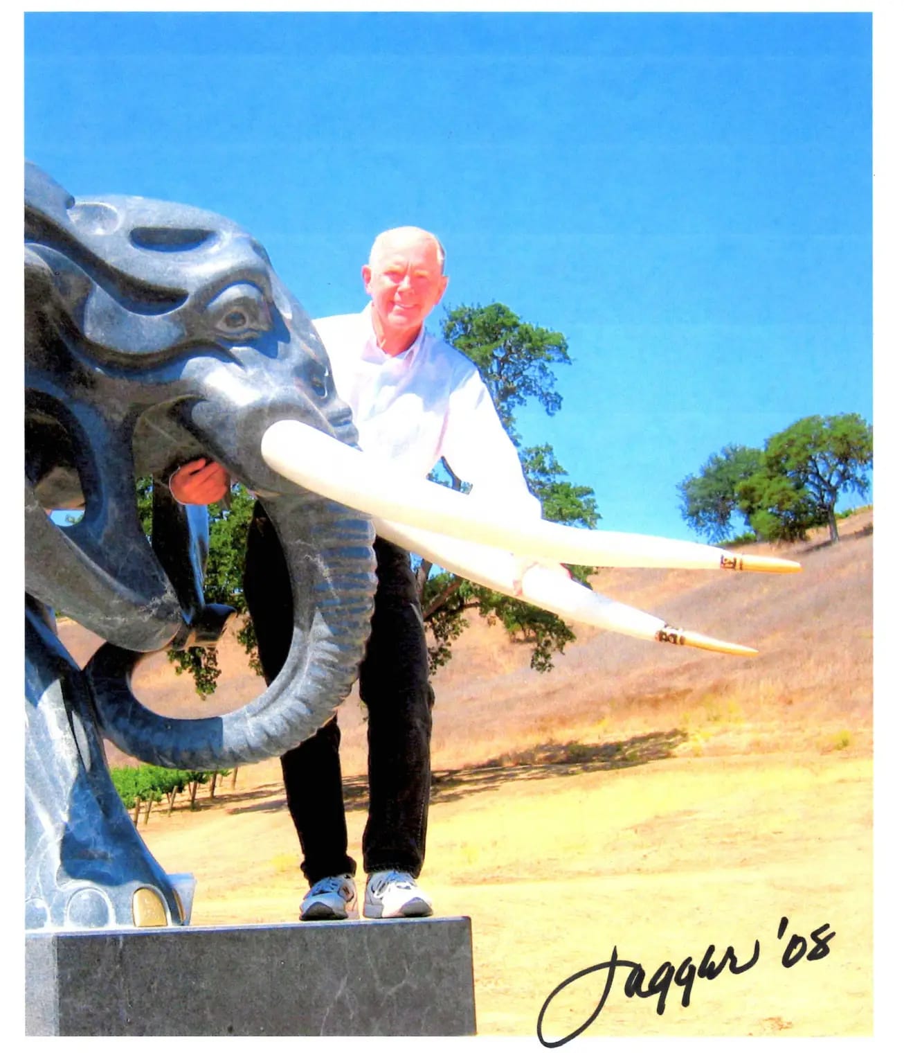 A photograph of a man standing behind the face of a grey stone elephant with long white tusks. He is holding the elephant's mouth. The photo is signed "Jagger '08" in the bottom right corner. 