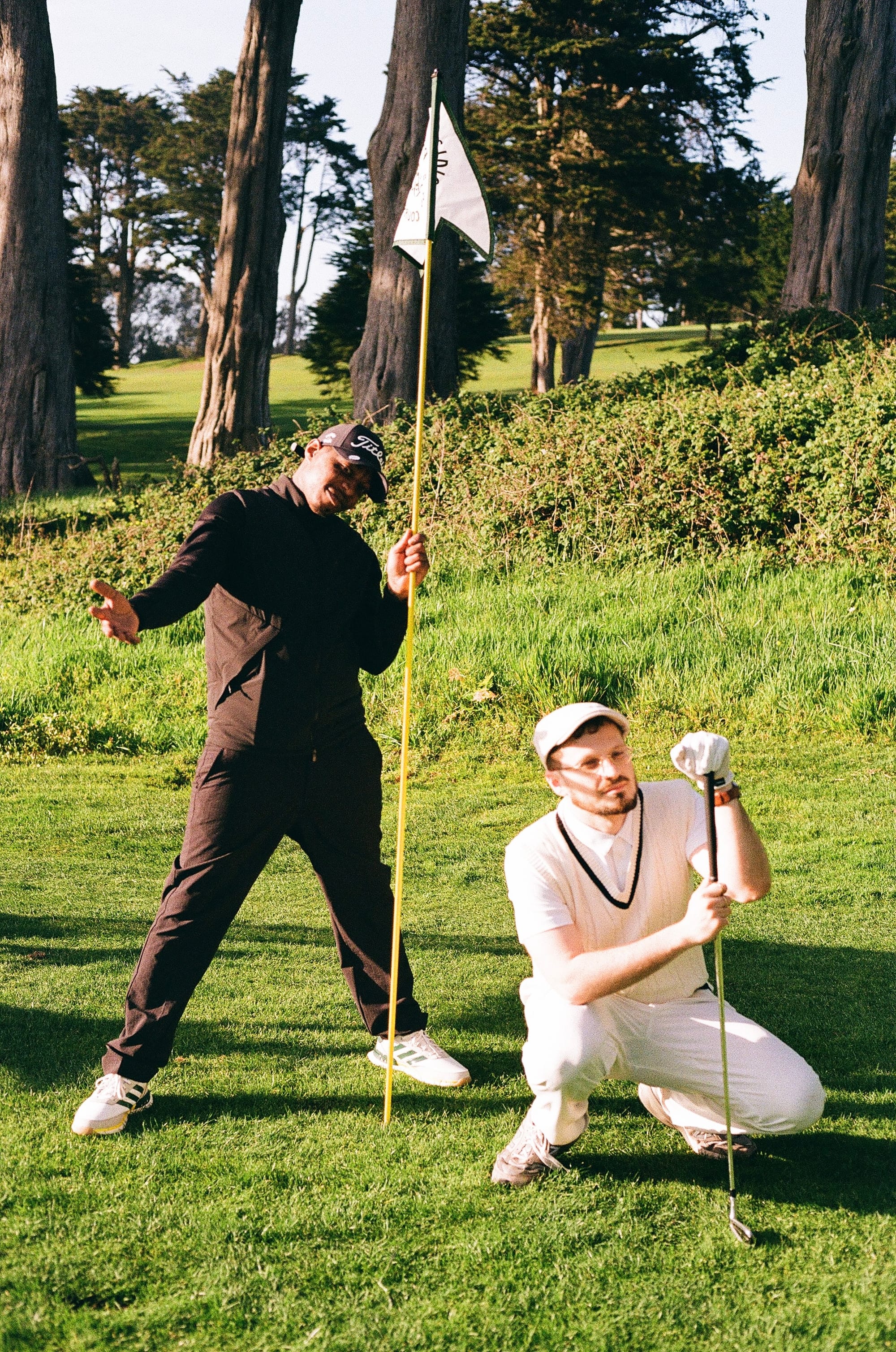 A man in black holds a golf flag on a golf course. A light-skinned man in white crouches down beside him while holding a golf club.
