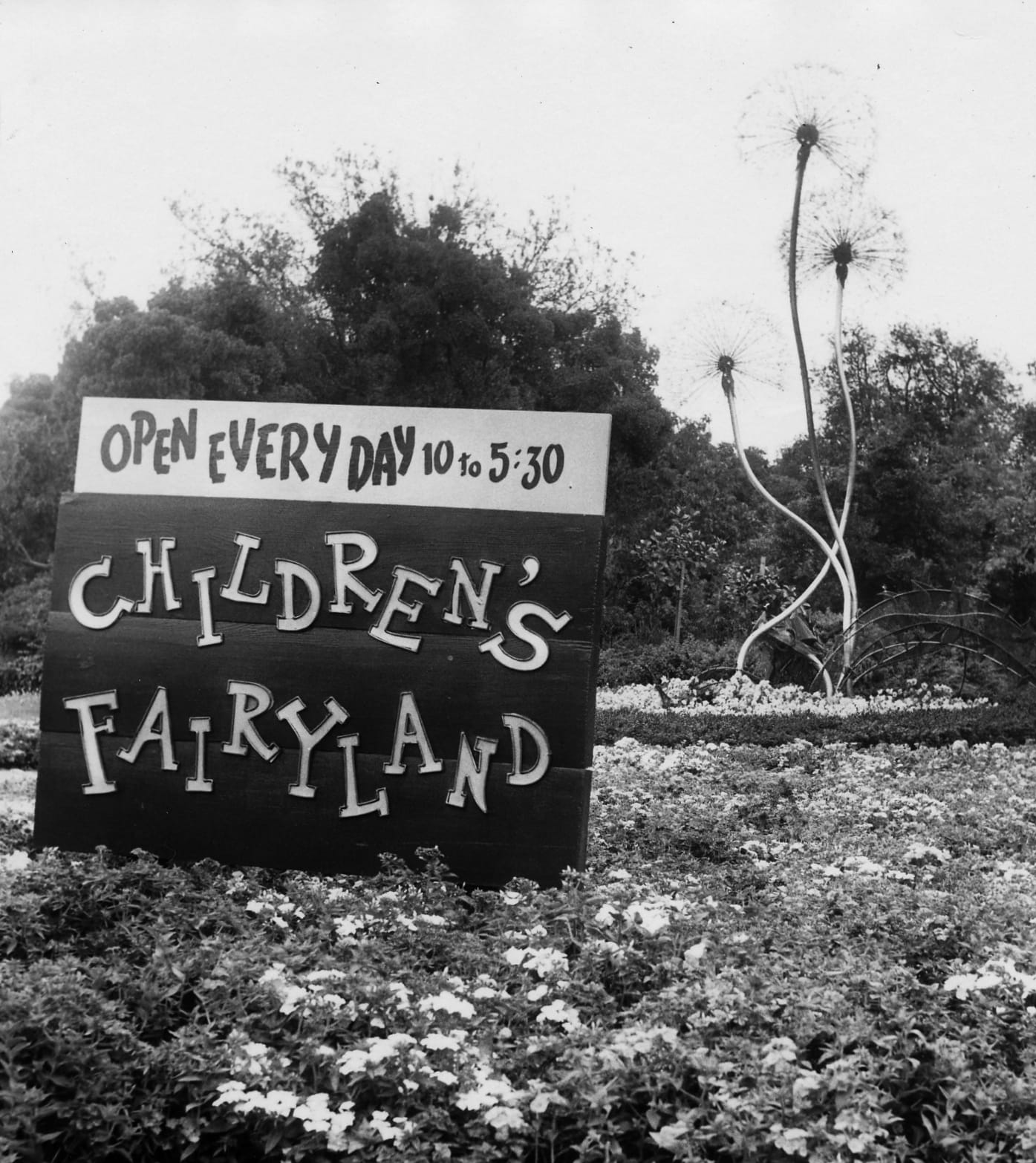 A black and white photograph of the front of Children's Fairyland, which shows the three tall metal dandelion puff sculptures and a wooden sign that says "Open Every Day 10 to 5:30 Children's Fairyland"