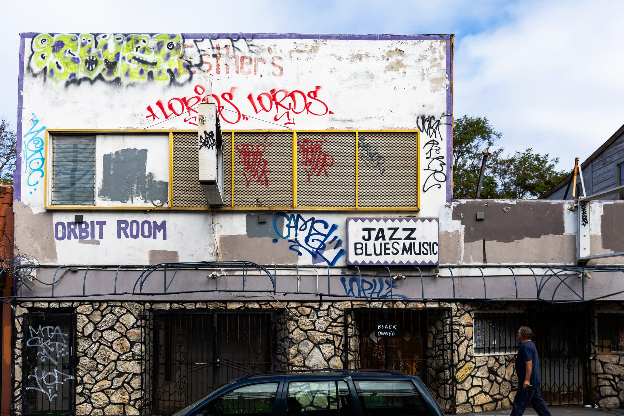 A building that has been painted with graffiti. The lower portion of the building has a faux stone wall, and the sign above the wall says "ORBIT ROOM" and "JAZZ BLUES MUSIC."