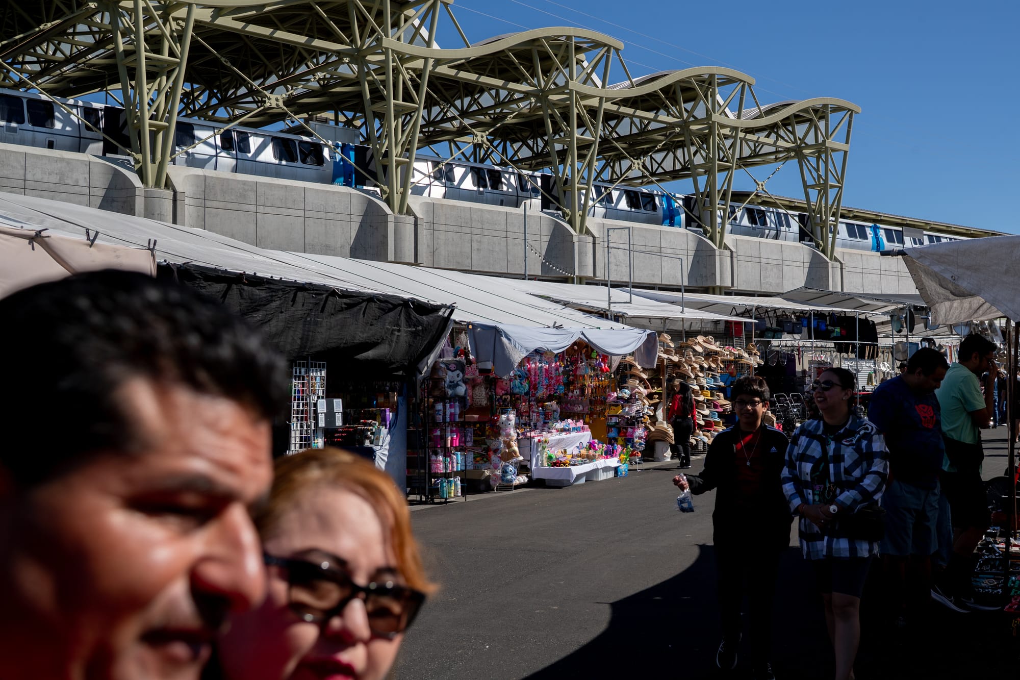 People walk through a flea market while a BART train arrives at the station above them.