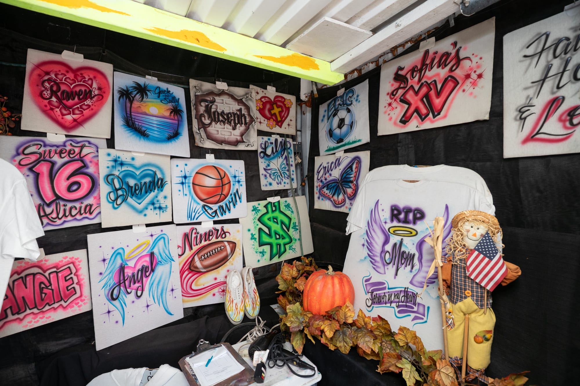 Airbrushed signs and a t-shirt on display with autumnal decor inside of a market stall.