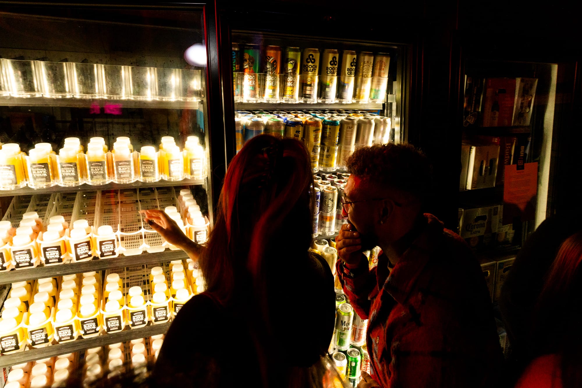 Two back lit people look a bottled cocktails in the convenience store fridge. 