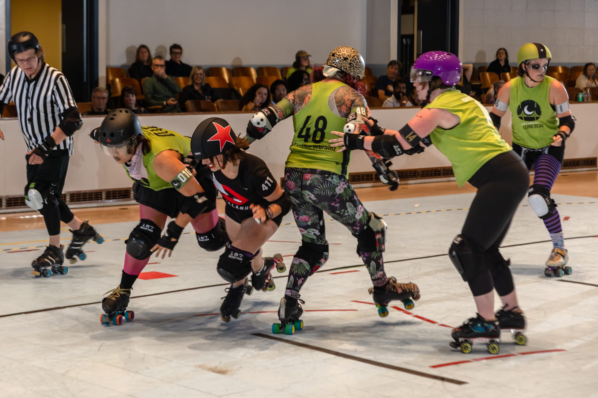 Four skaters in green jerseys are trying to stop a skater in a black outfit with a big red star on their helmet. The skater with the star is pushing through them anyway.