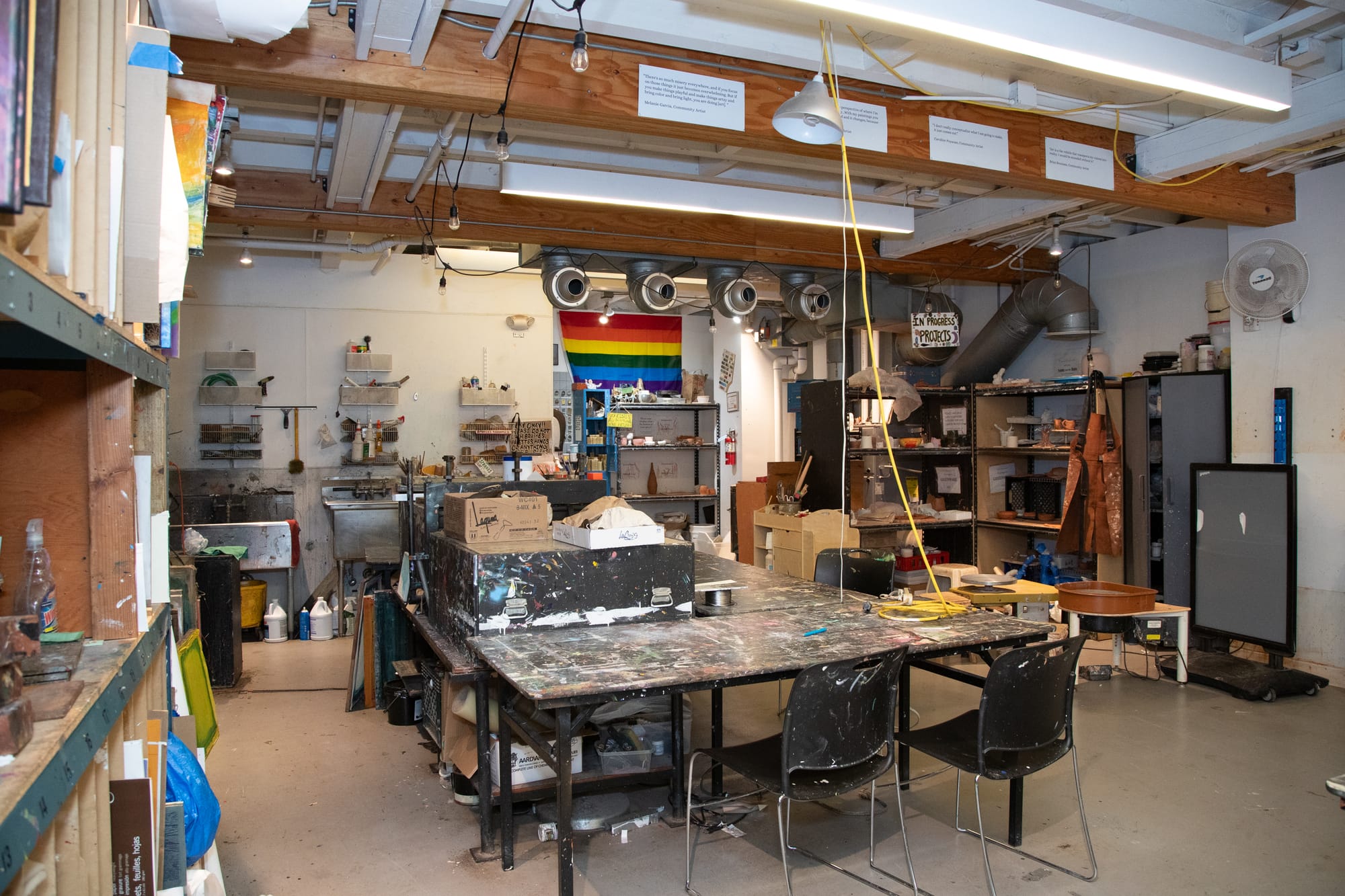 A large table in the center of an art studio room is covered in paint. Two empty black chairs sit before it.