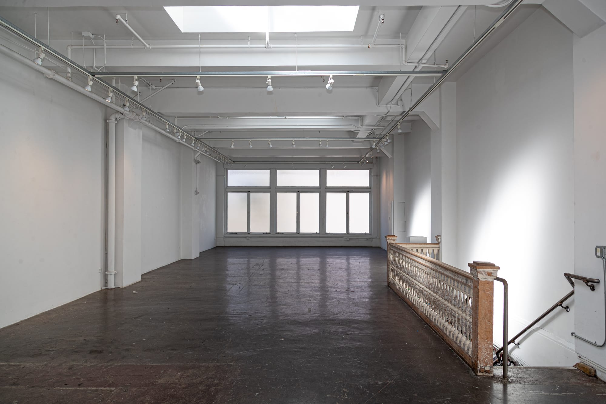 A large empty white room with stairs going down on the right-hand side. The floor is dark and there is gallery-style lighting on the ceilings.