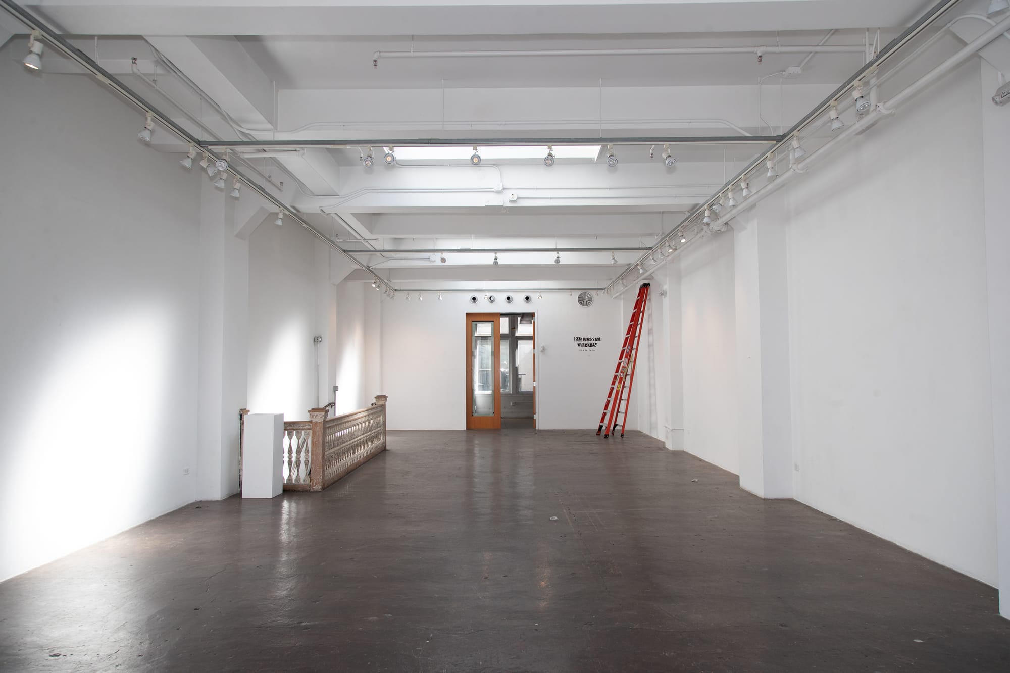 A large empty white room has a red ladder leaning against a wall in the back. The floor is dark and there is gallery-style lighting on the ceilings.