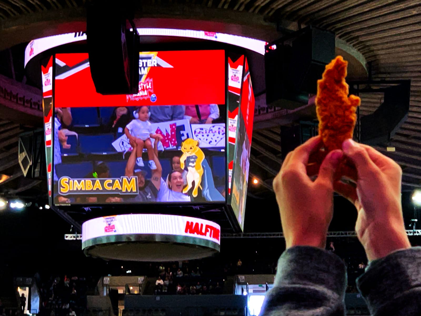 A child is held by a man on the jumbotron while a child in the audience holds up a chicken tender. 