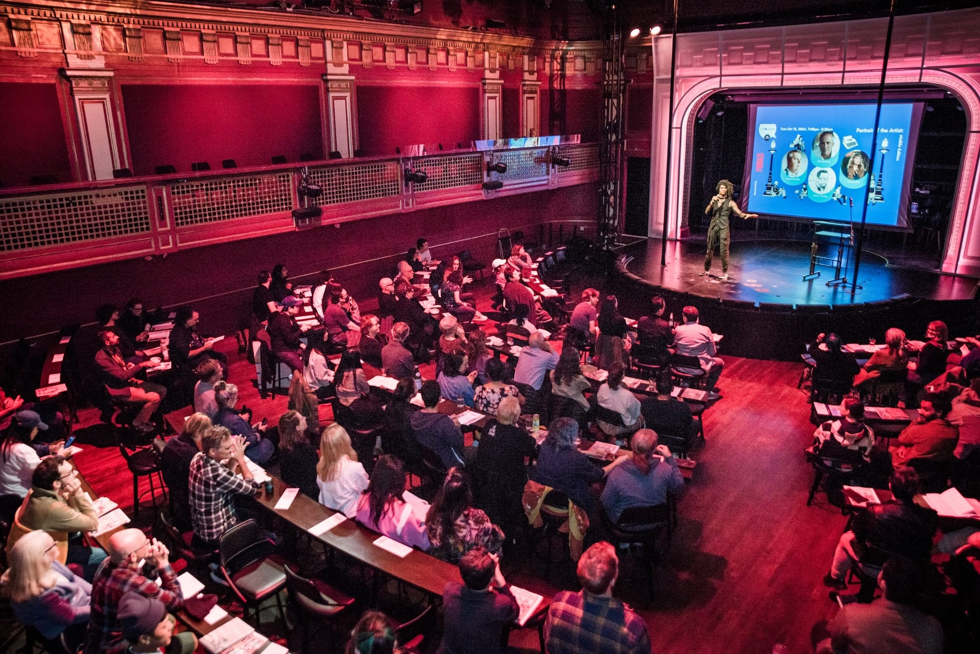 a packed theater with a person on stage reading