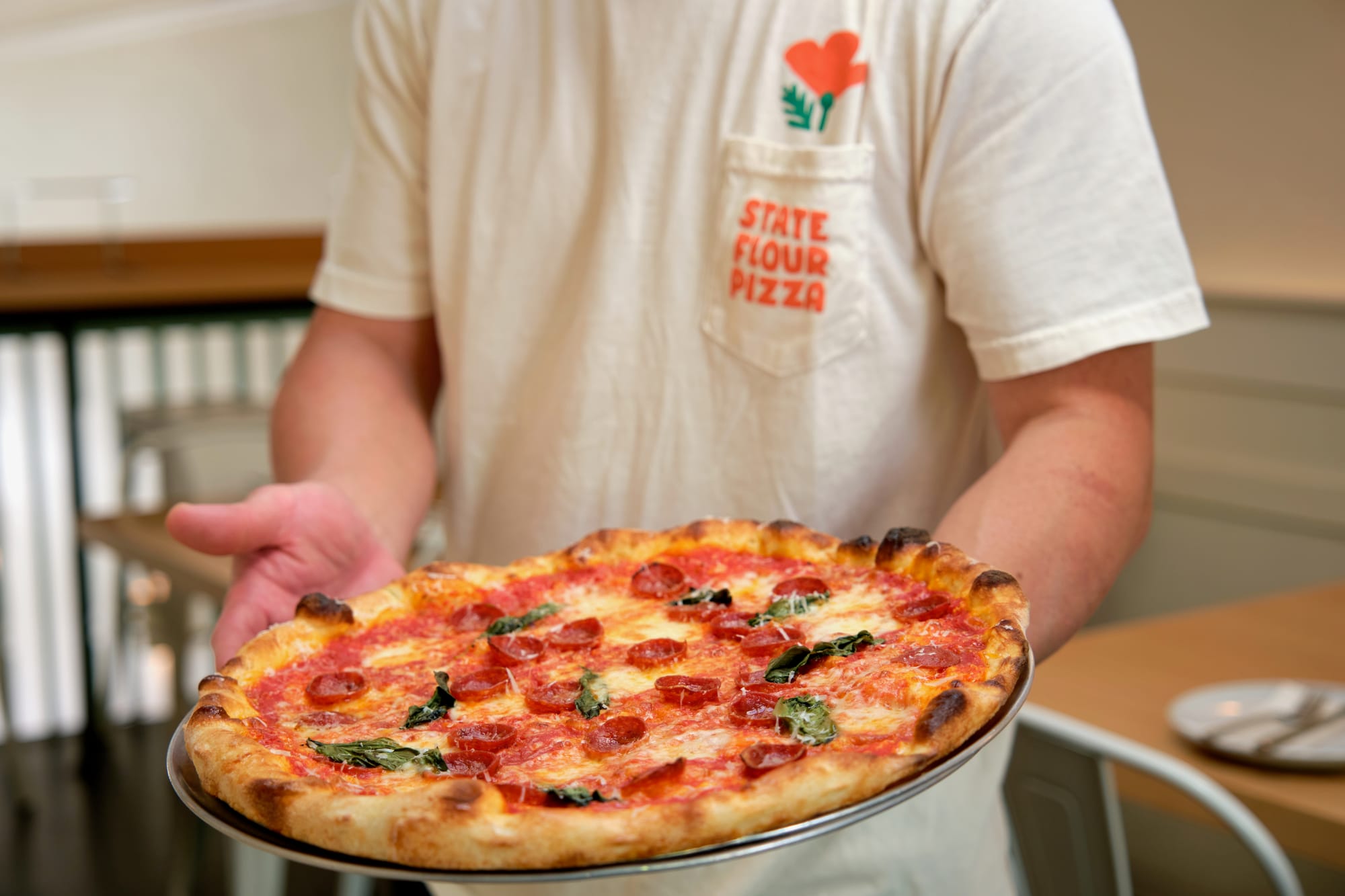 A person in a t-shirt holds a margherita and pepperoni pizza inside of a restaurant.