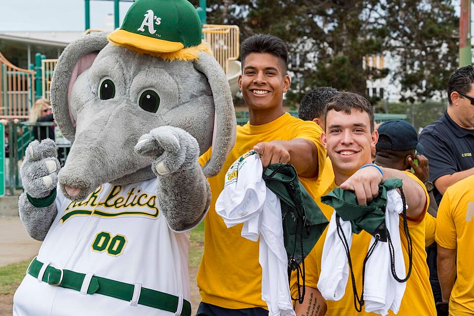 On the right, two athletes pointing at the camera and smiling. On the left, an elephant mascot wearing a green hat with a yellow brim and an A's logo on it.
