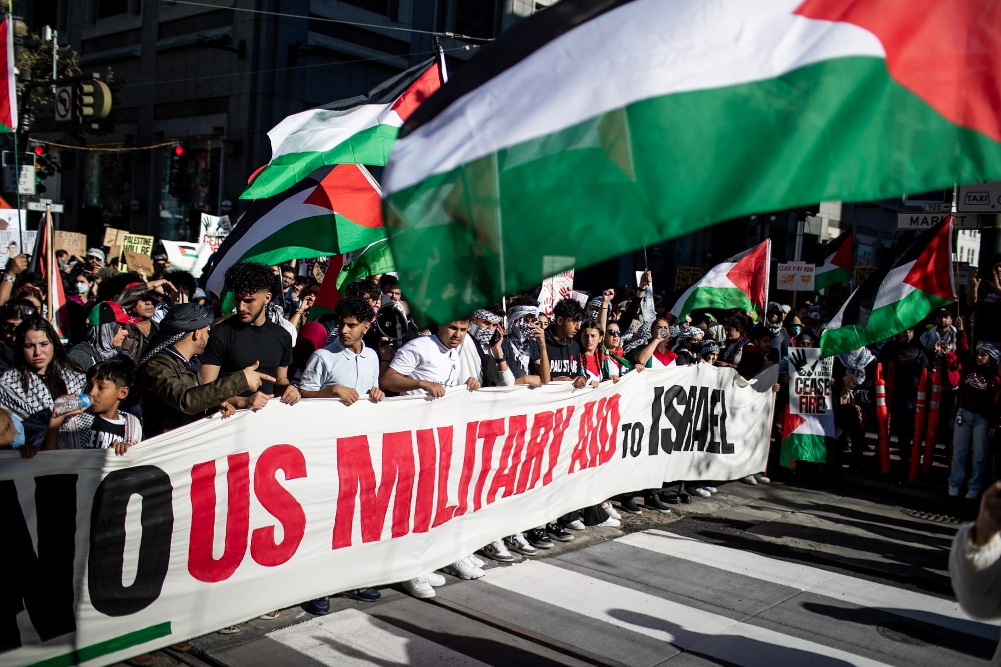 Thousands of protesters march down a street holding a large banner and  Palestinian flags. 