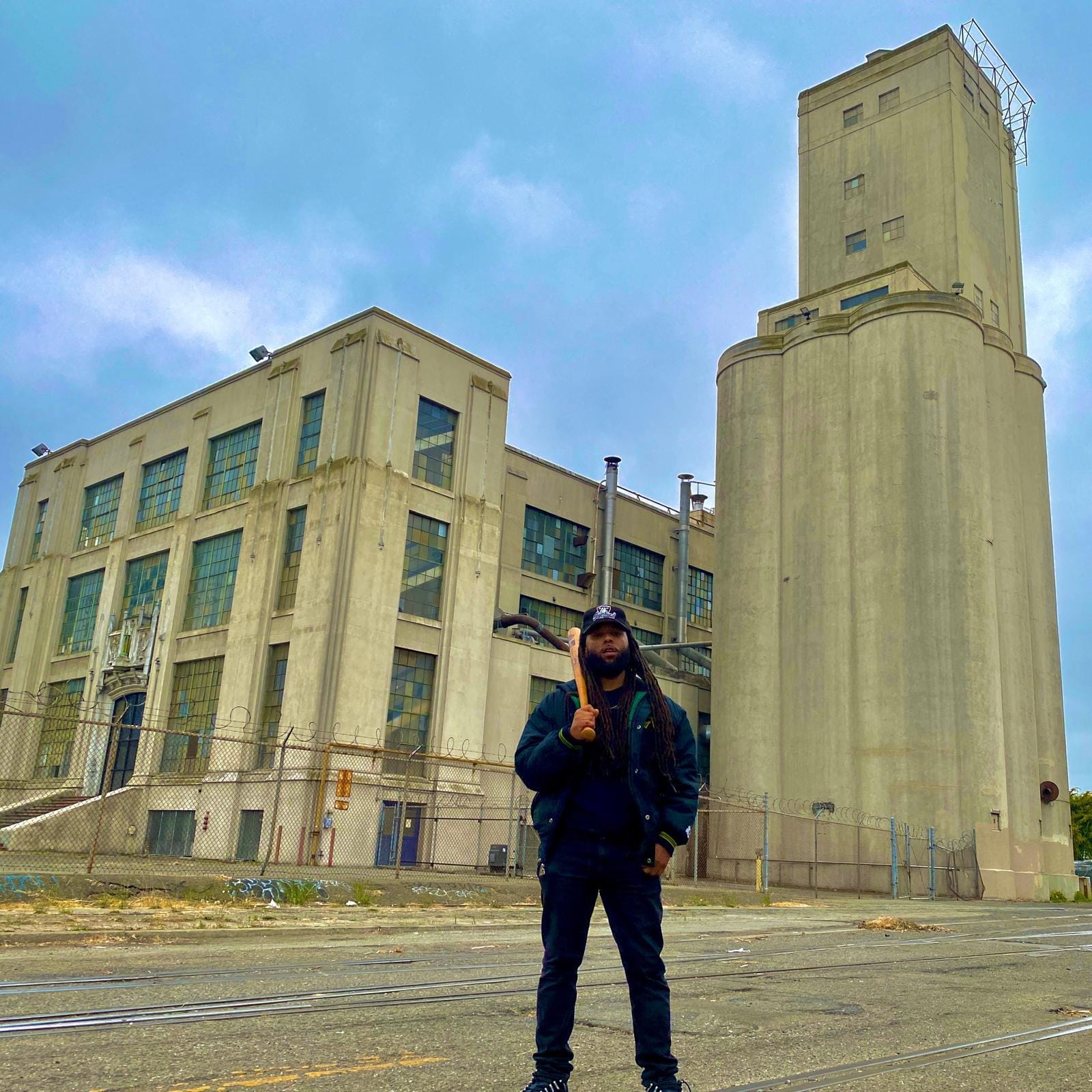 a young Black man with braids stands in front of a concrete skyscraper holding a tool 