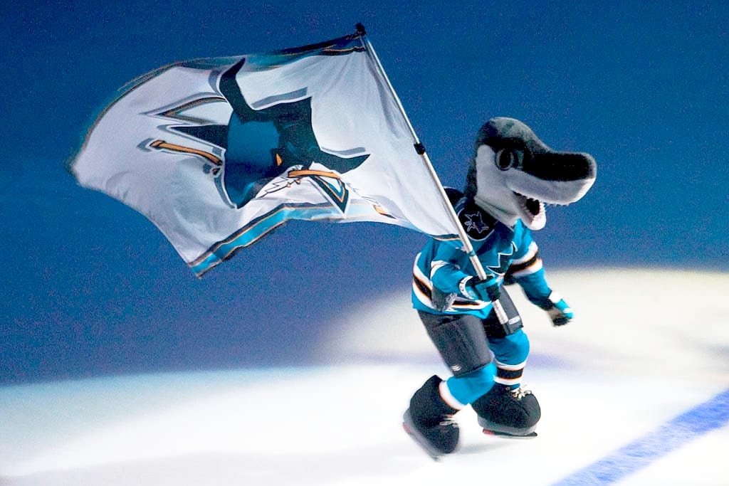 A shark mascot skating on the ice, holding a flag with the San Jose Sharks logo on it.
