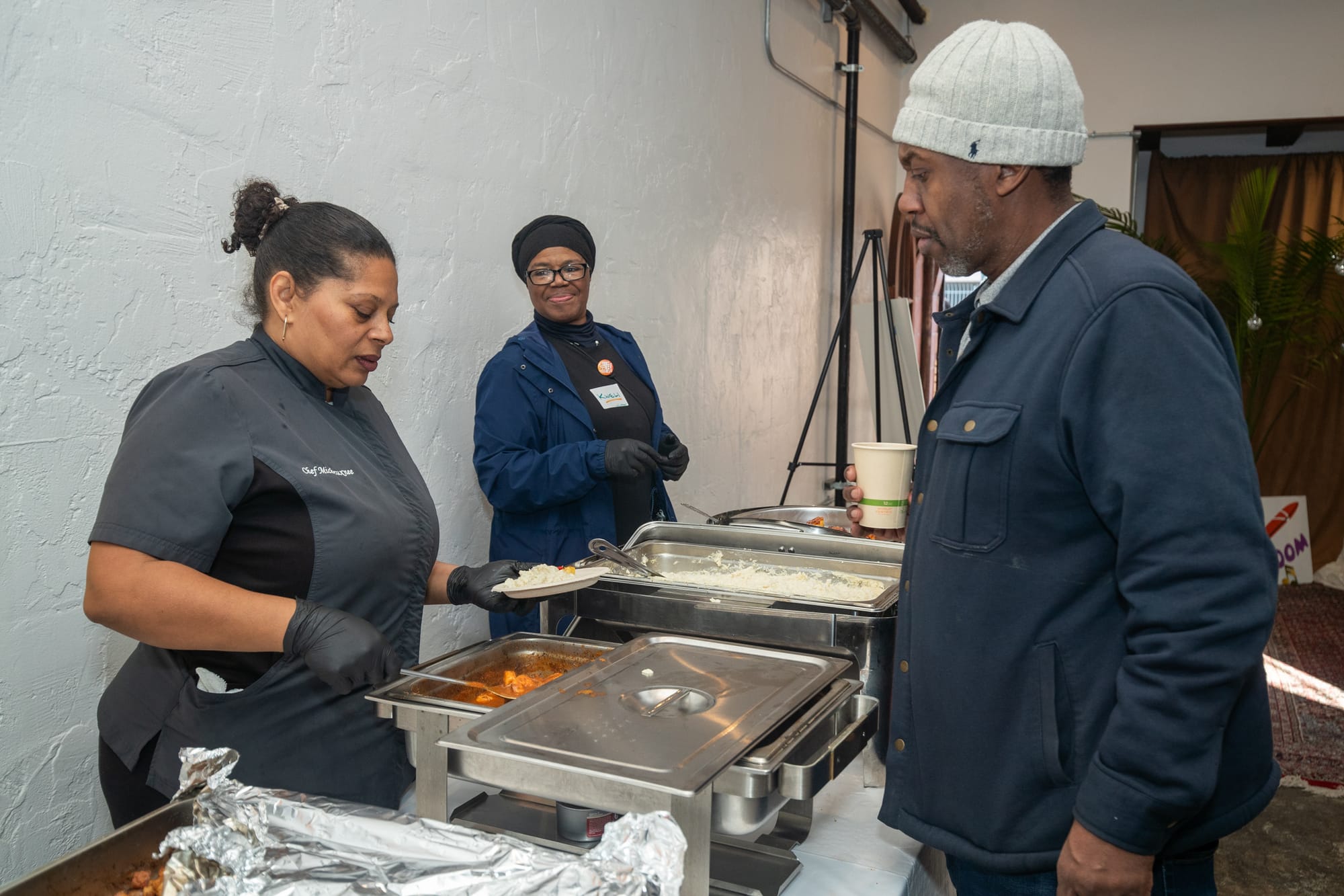 A Black woman wearing a black chef coat serves food from a metal steam table tray.