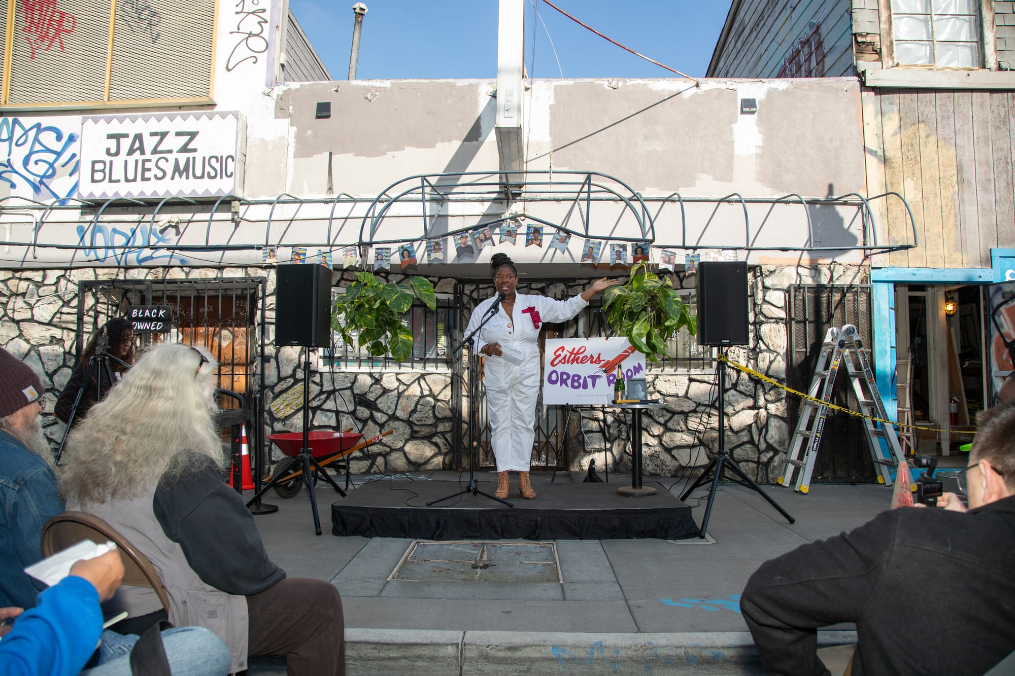 A Black woman in a white jumpsuit stands on a small stage and addresses the crowd. Behind her is a faux stone facade, caution tape, a ladder and a wheelbarrow.