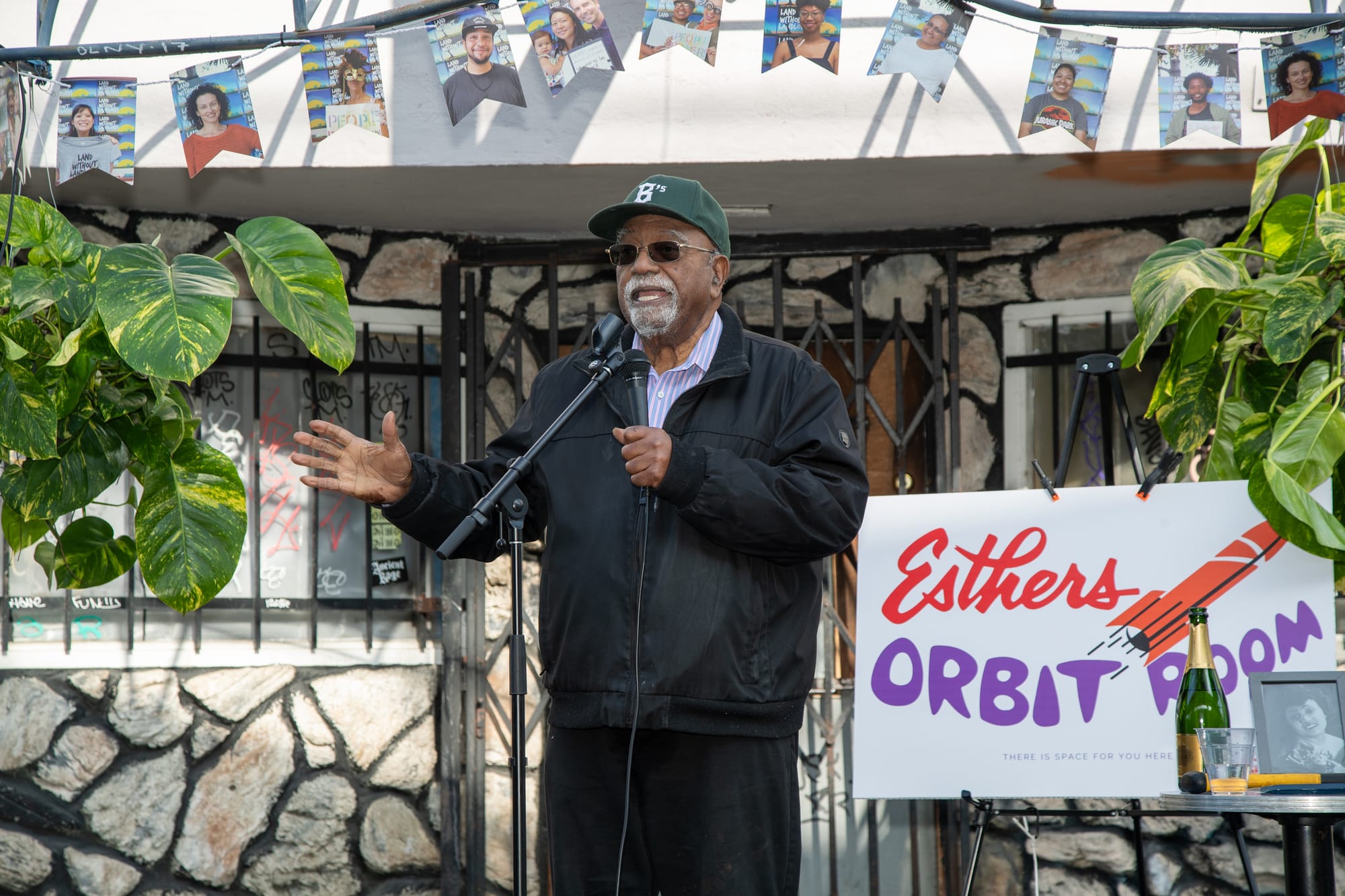 A Black man with a white goatee holds a microphone and speaks into it. Behind him is a sign for Esthers Orbit Room, and to the right and left are hanging plants.