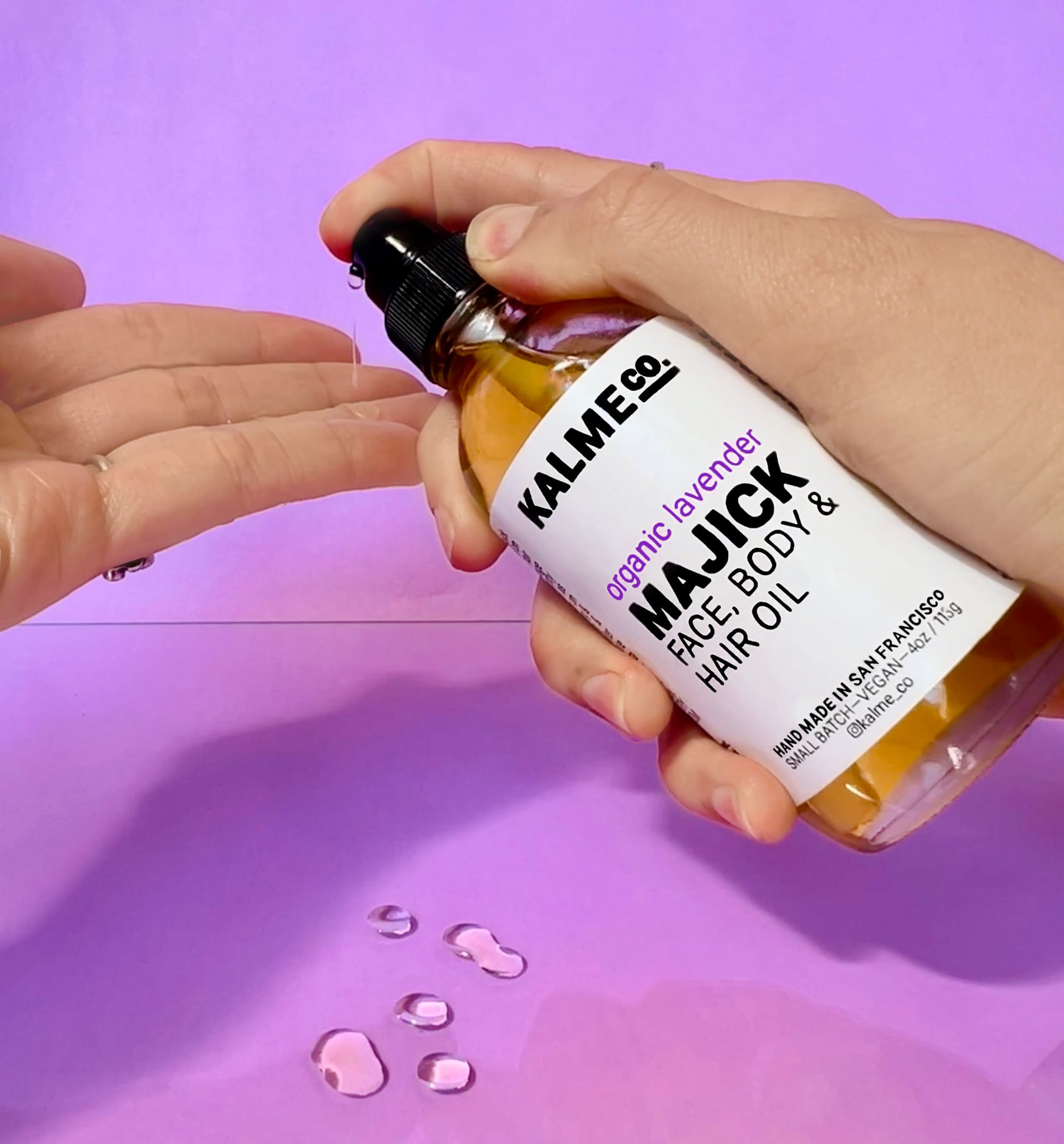 a purple background with hands pumping from a bottle that reads 'Kalme Co. organic lavender majick face, body & hair oil, hand made in San Francisco'