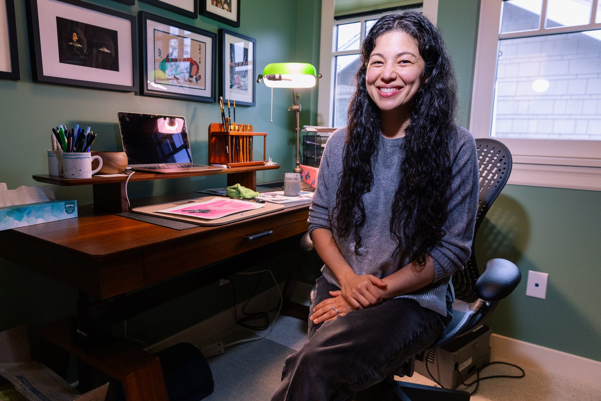 A woman sitting in front of a desk. The woman is smiling, and has long, wavy dark hair. She's wearing a blue sweater. 