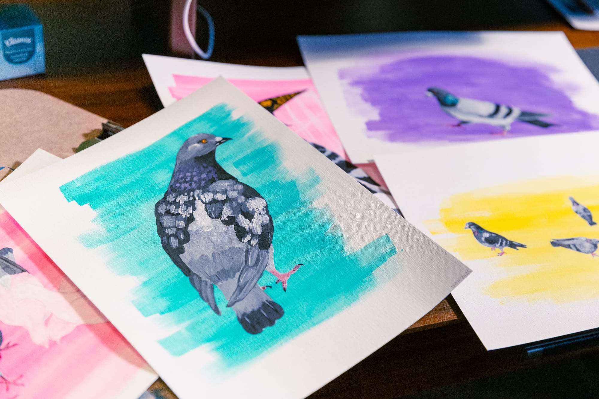 A painting of a pigeon, with a teal wash background. Nearby, out of focus, more paintings of pigeons with different colored backgrounds (purple, yellow, pink).