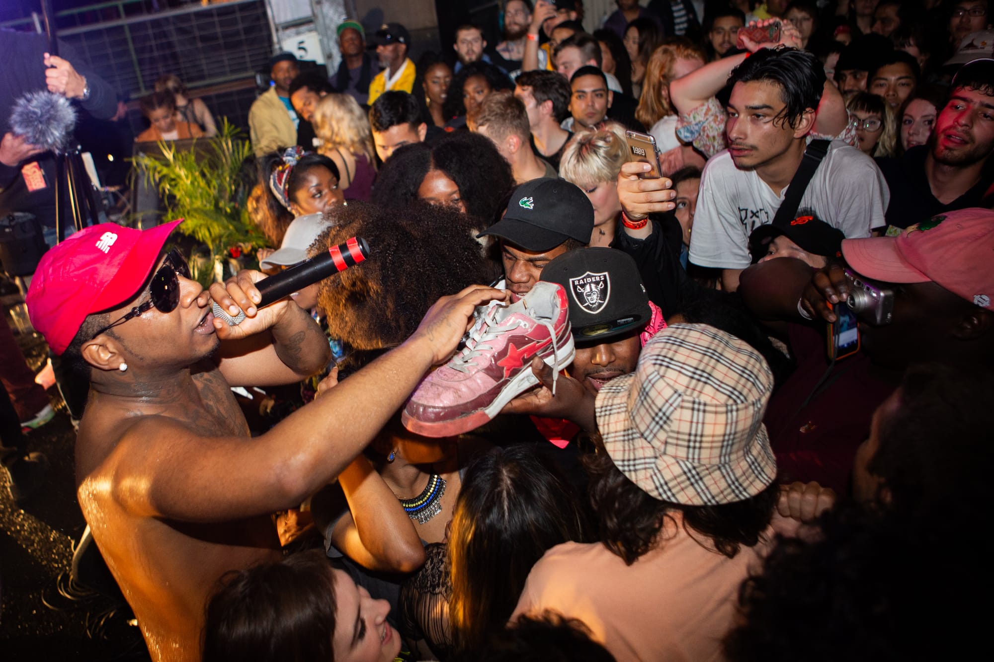 a young man in a red ball cap and sunglasses holds a shoe while performing at a crowded party