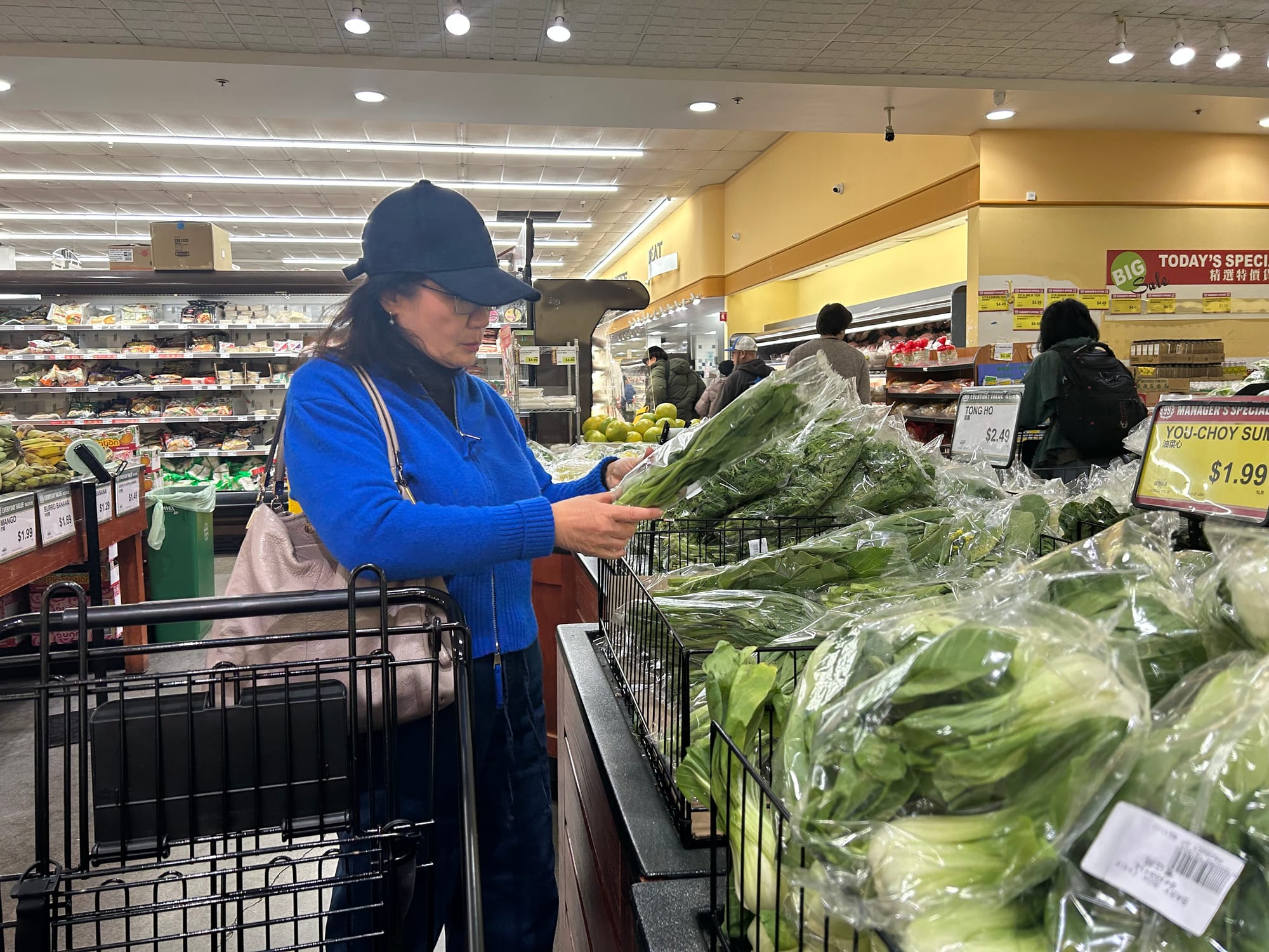 A woman in a blue sweater picking up and examining a plastic-wrapped bundle of Asian greens in a grocery store.