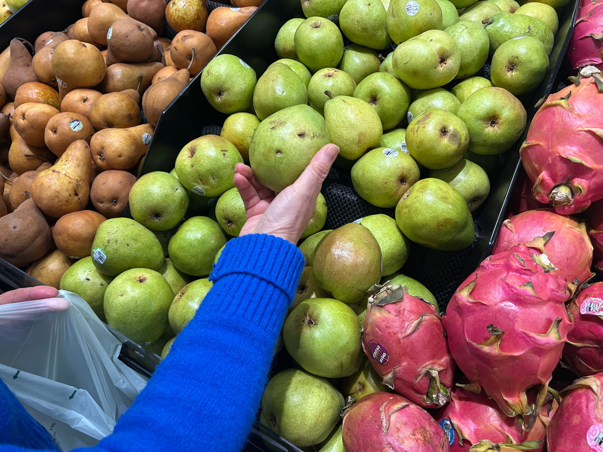 A person wearing a blue sweater picks up a green pear from a display of fruits including brown and green pears and pink dragonfruits.