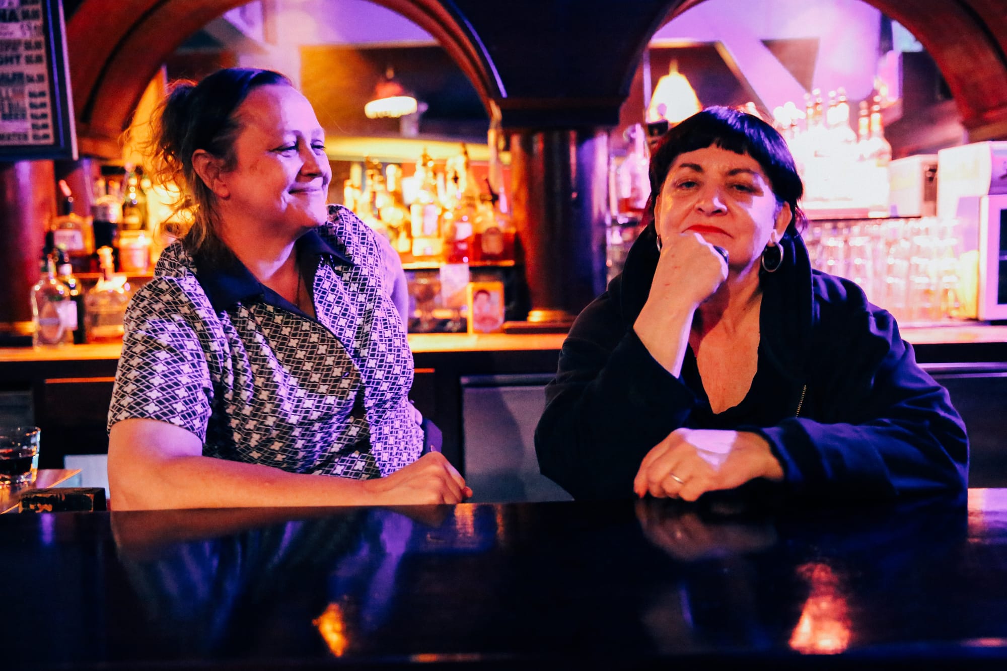 two women pose behind a bar in a club, one smiling at the other, the other looking at the camera