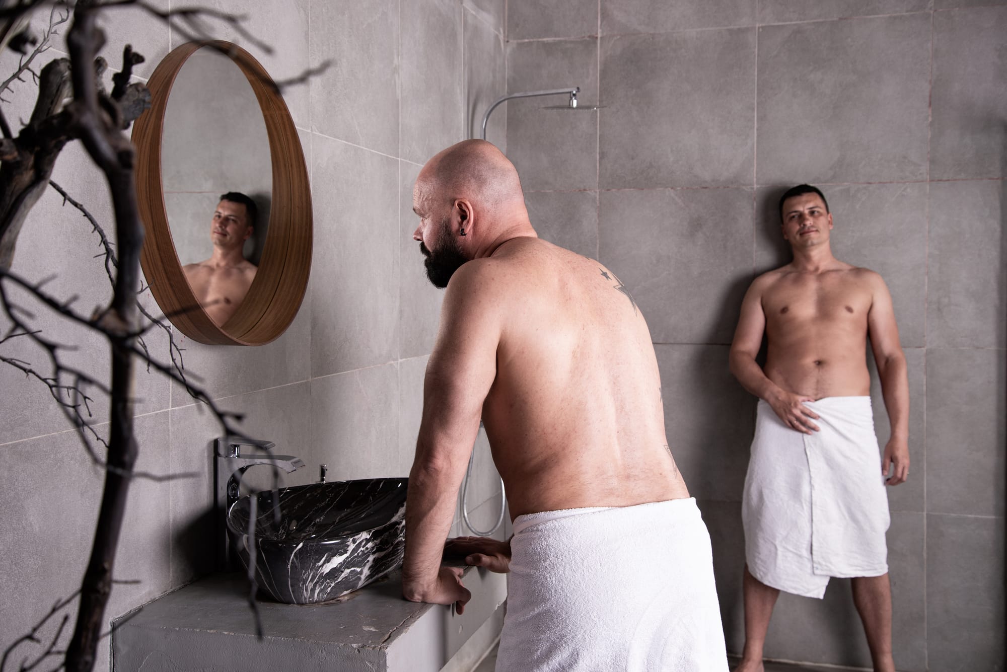 Two men in a bathroom wearing only towels. One is looking at the other with a solicitous expression.