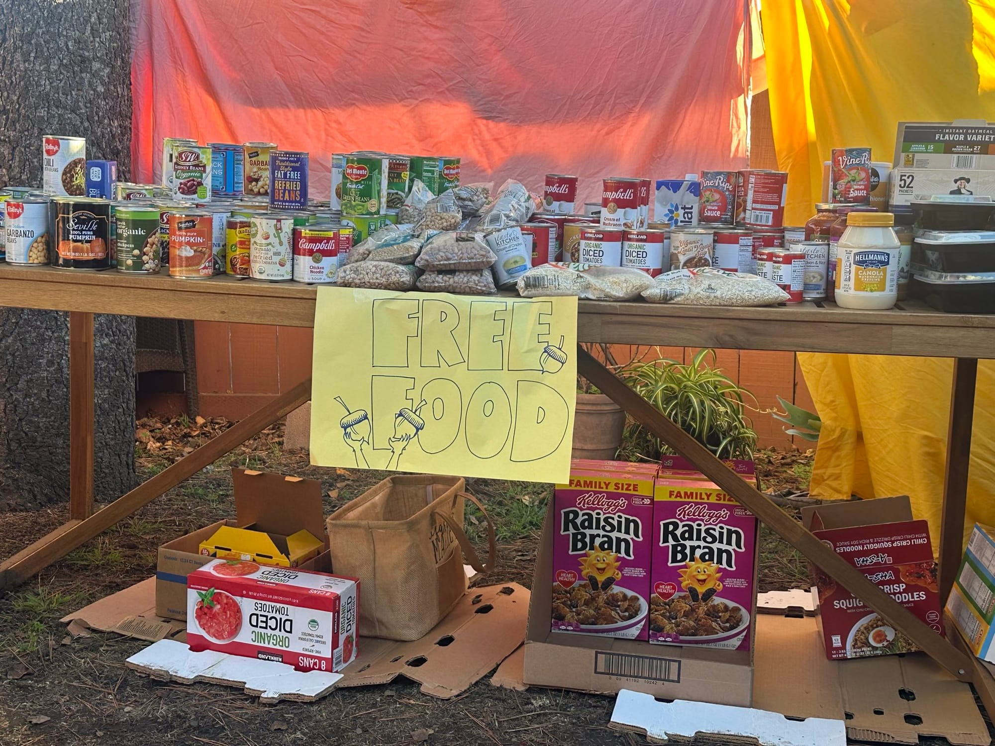 A table covered in mostly nonperishable food like cans, beans, and rice, but also boxes of cereal, vegan mayonnaise and instant oats. A sign on the table says "FREE FOOD"