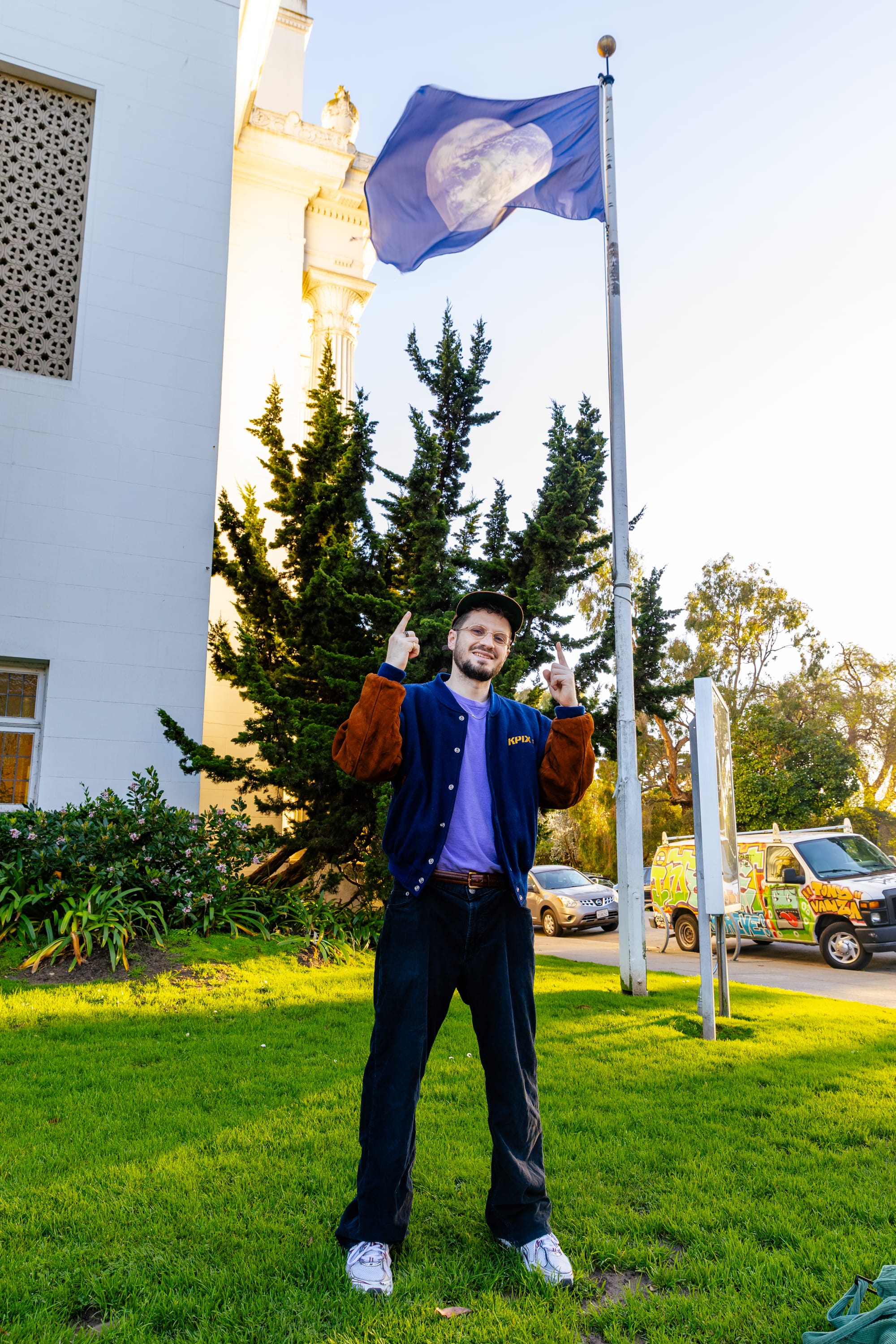 A rapper stands beneath a flag of the globe in front of a building and points up at it while smiling.