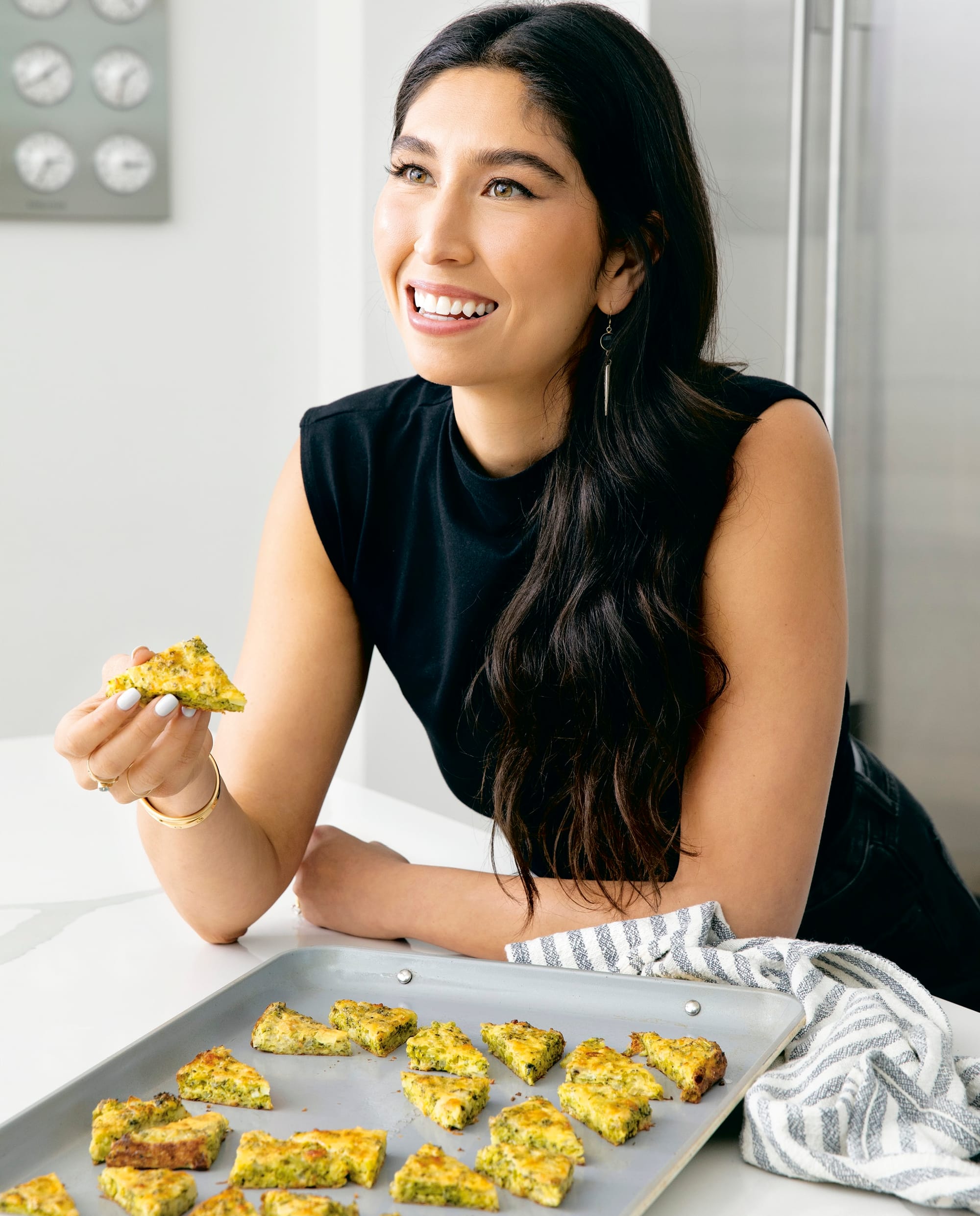 A woman with long black hair holds a piece of frittata. She's smiling at something out-of-frame.