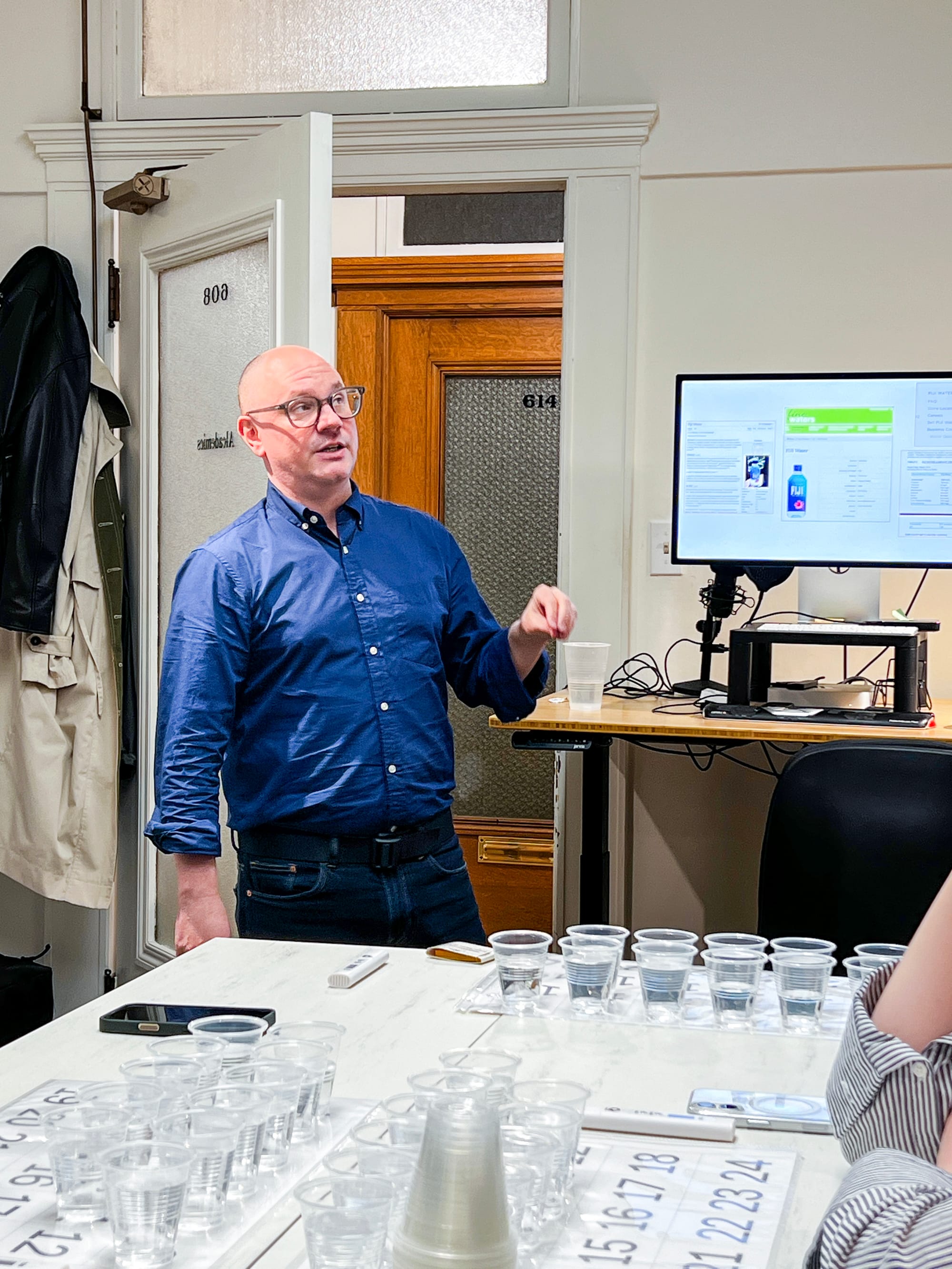 a man in ablue button down shirt and jeans stands speaking at a table full of plastic cups filled with water