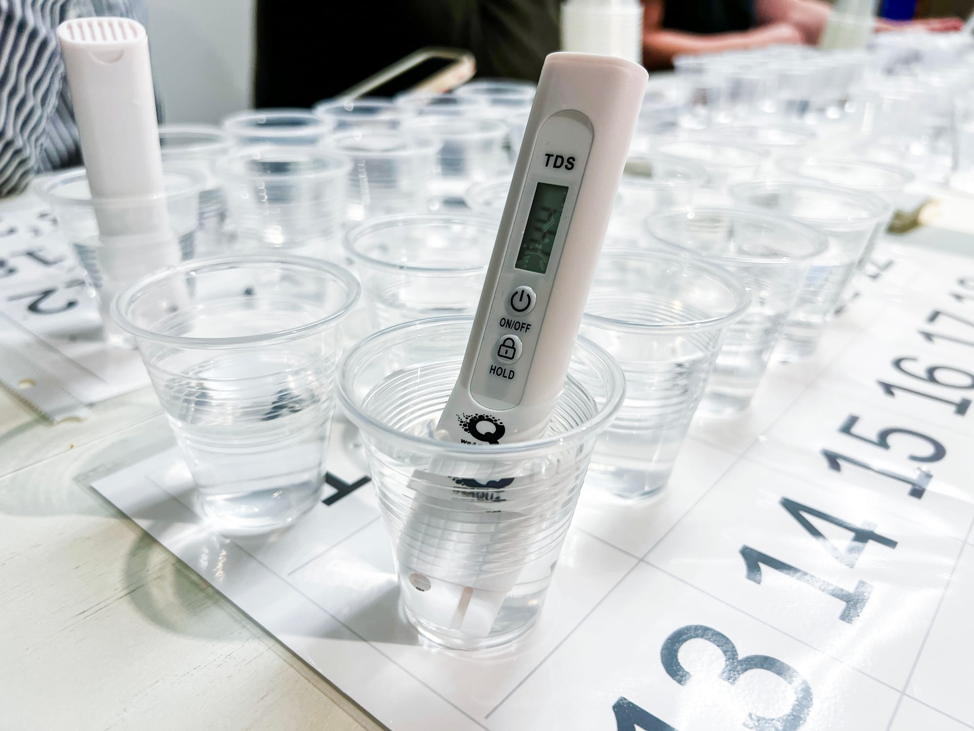 a thermometer type device with 'TDS' on it is submerged in a plastic cup of water next to other plastic cups of water on a table