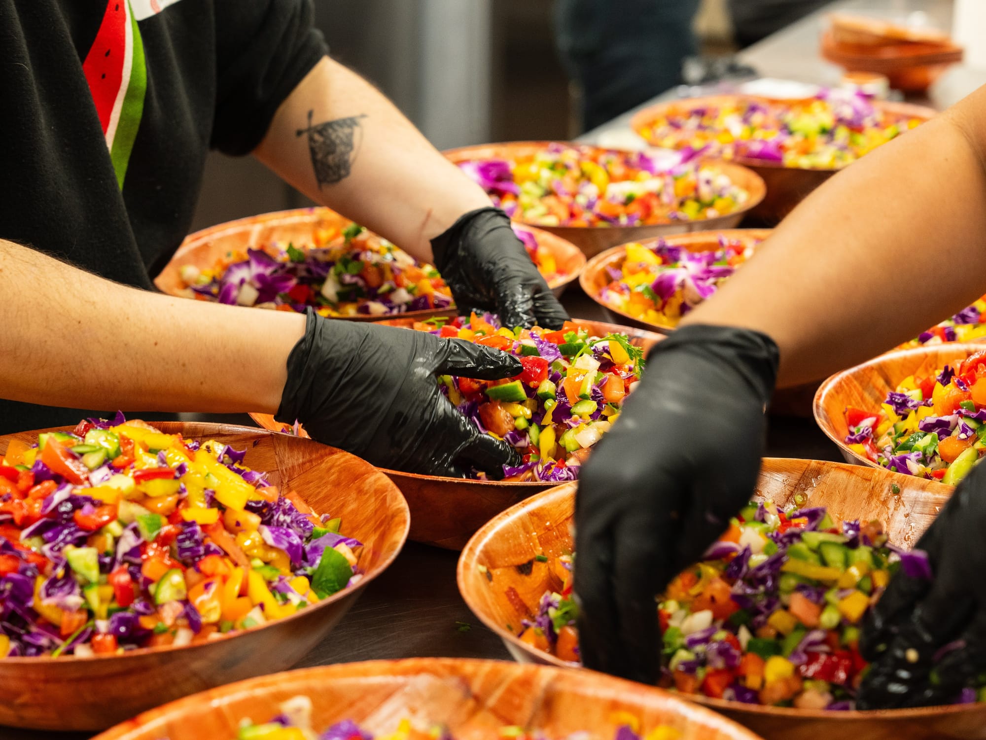 A close-up of hands wearing black gloves mixing colorful salads in wooden bowls.