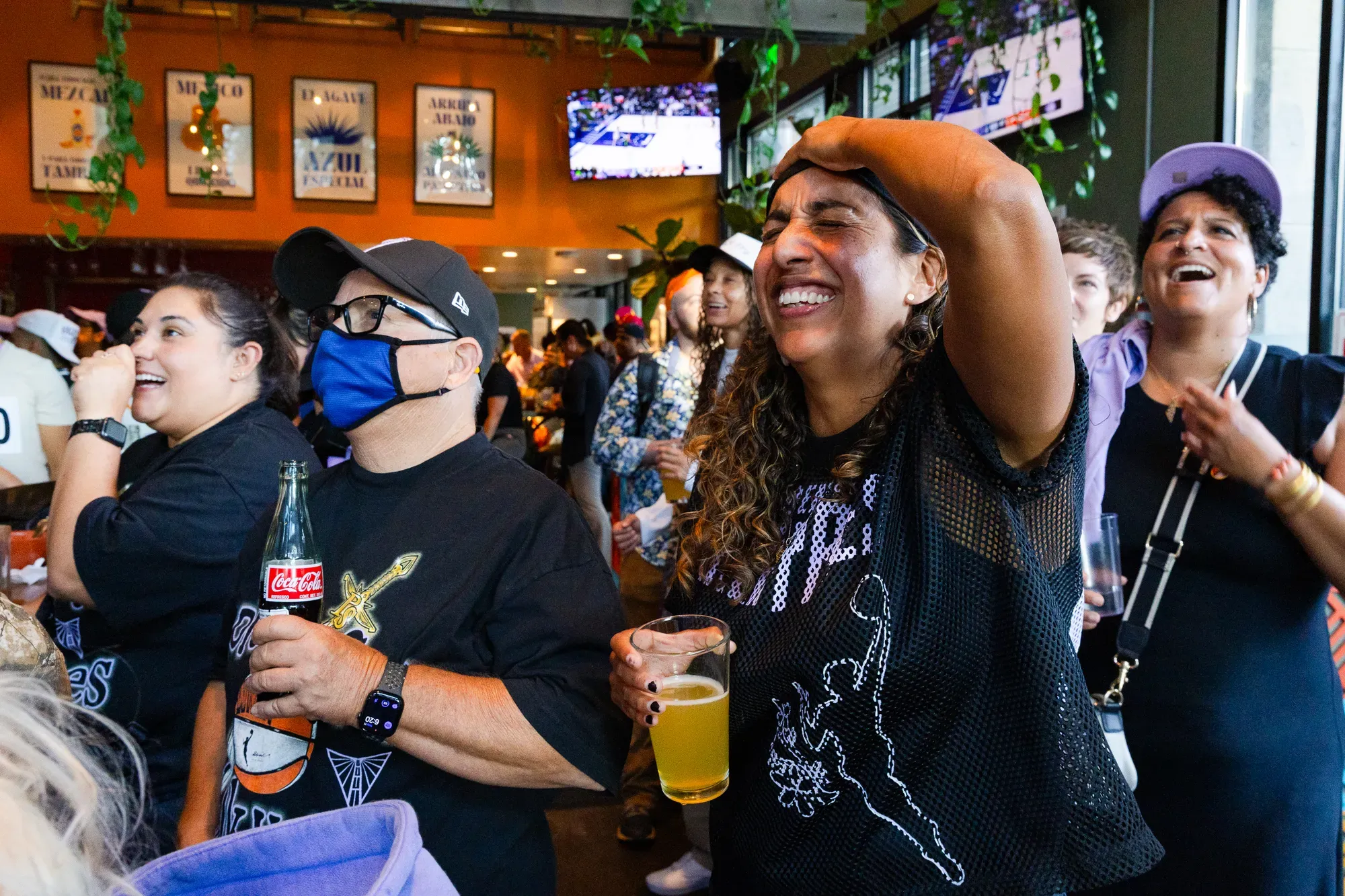a group of people in lavender and black sports jerseys grimace while drinking beers and sodas and looking up at a sports bar