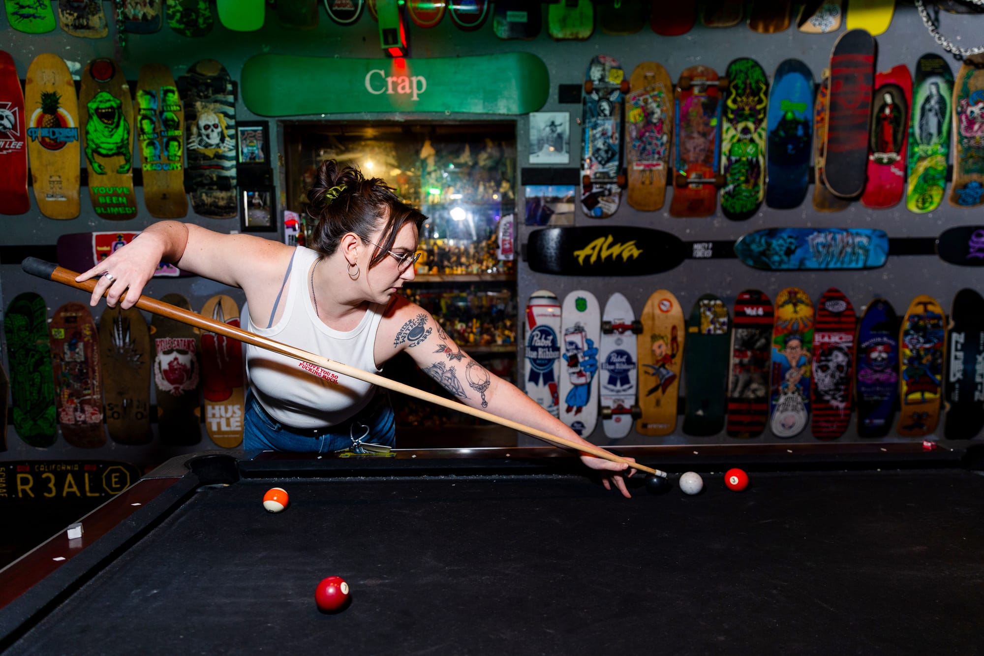 a person in a white tank top and jeans is about to make a shot with white and red balls on a pool table at a bar with skateboards on the wall in the background
